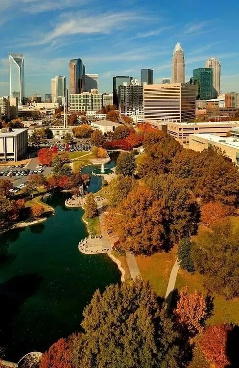 A cityscape with a park in the foreground, featuring a pond, trees with fall foliage, and a fountain, skyscrapers in the background, and a partly cloudy blue sky.