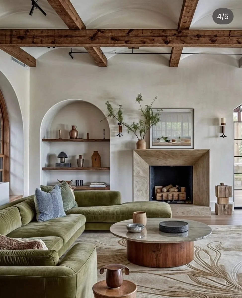 Living room with a green velvet sectional sofa, a wooden round coffee table, a fireplace with wood logs, built-in shelves with decorative vases, a large window, and ceiling wooden beams.