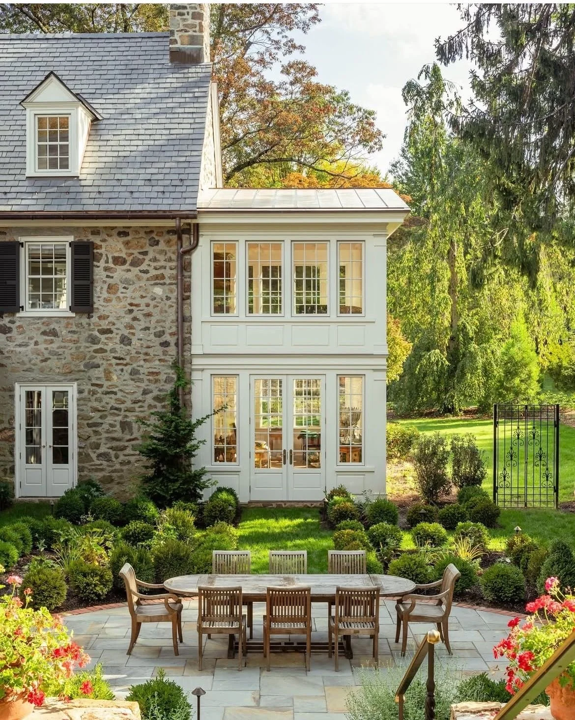 Backyard view of a stone house with large porch windows, surrounded by green lawn, bushes, and trees, with a patio table and chairs in the foreground.