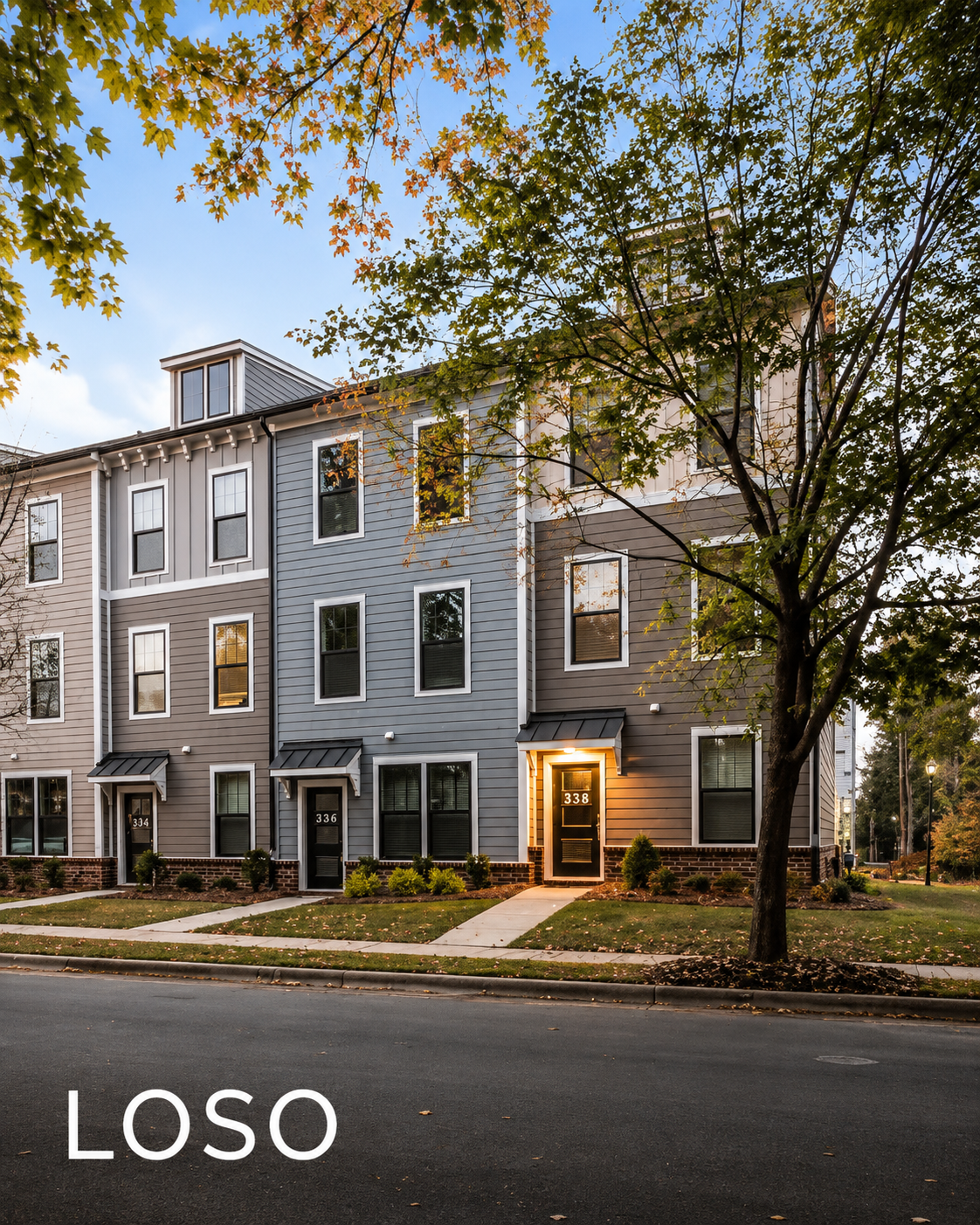 A modern multi-story apartment building with gray and beige siding, multiple windows, and front doors numbered 334, 336, and 338, with landscaping and a large tree in front. The building is illuminated by outdoor lighting, and trees with autumn leaves are visible in the foreground and background.