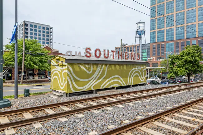 A yellow kiosk with white abstract patterns along a railroad track in an urban area, with buildings, trees, and a water tower in the background. The sign on the kiosk reads "SOUTHEND".