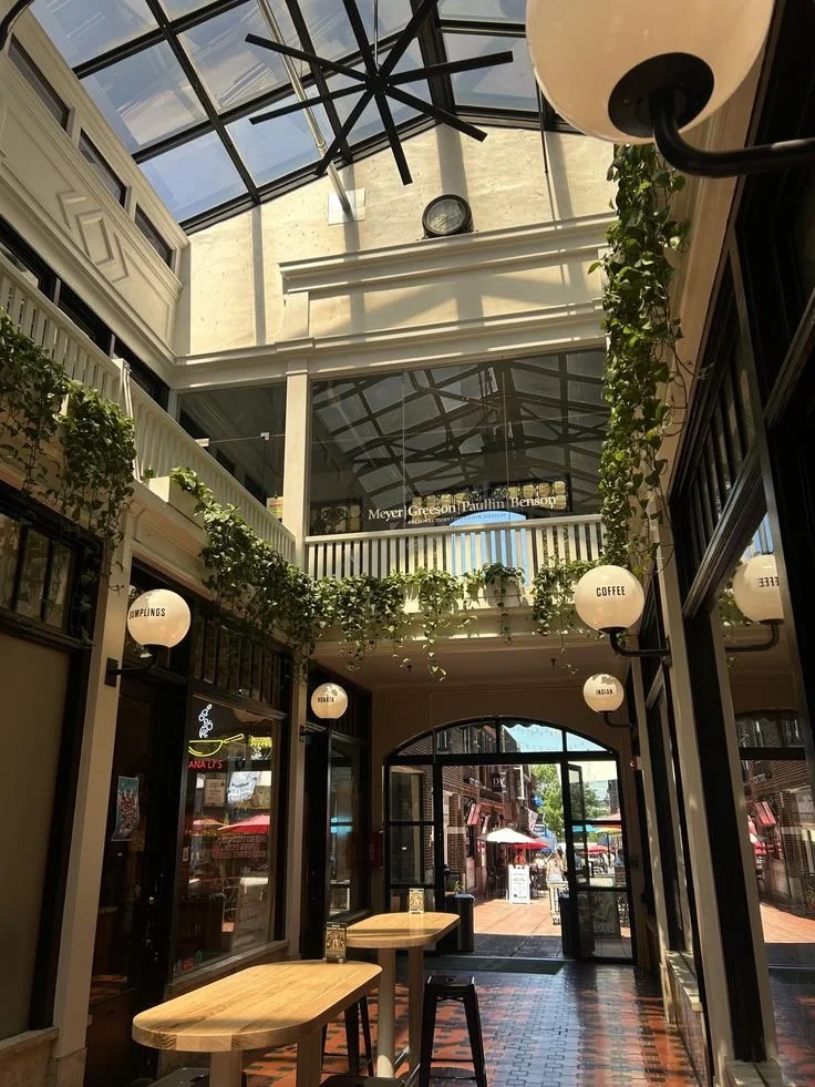 Interior of a bright, indoor shopping or dining area with a glass ceiling. There are hanging lights labeled with words like "COFFEE," "MUMPLINGS," and "ASIAN." There are green vines decorating the space, wooden tables and stools, and a view of an outdoor shopping area through glass doors.