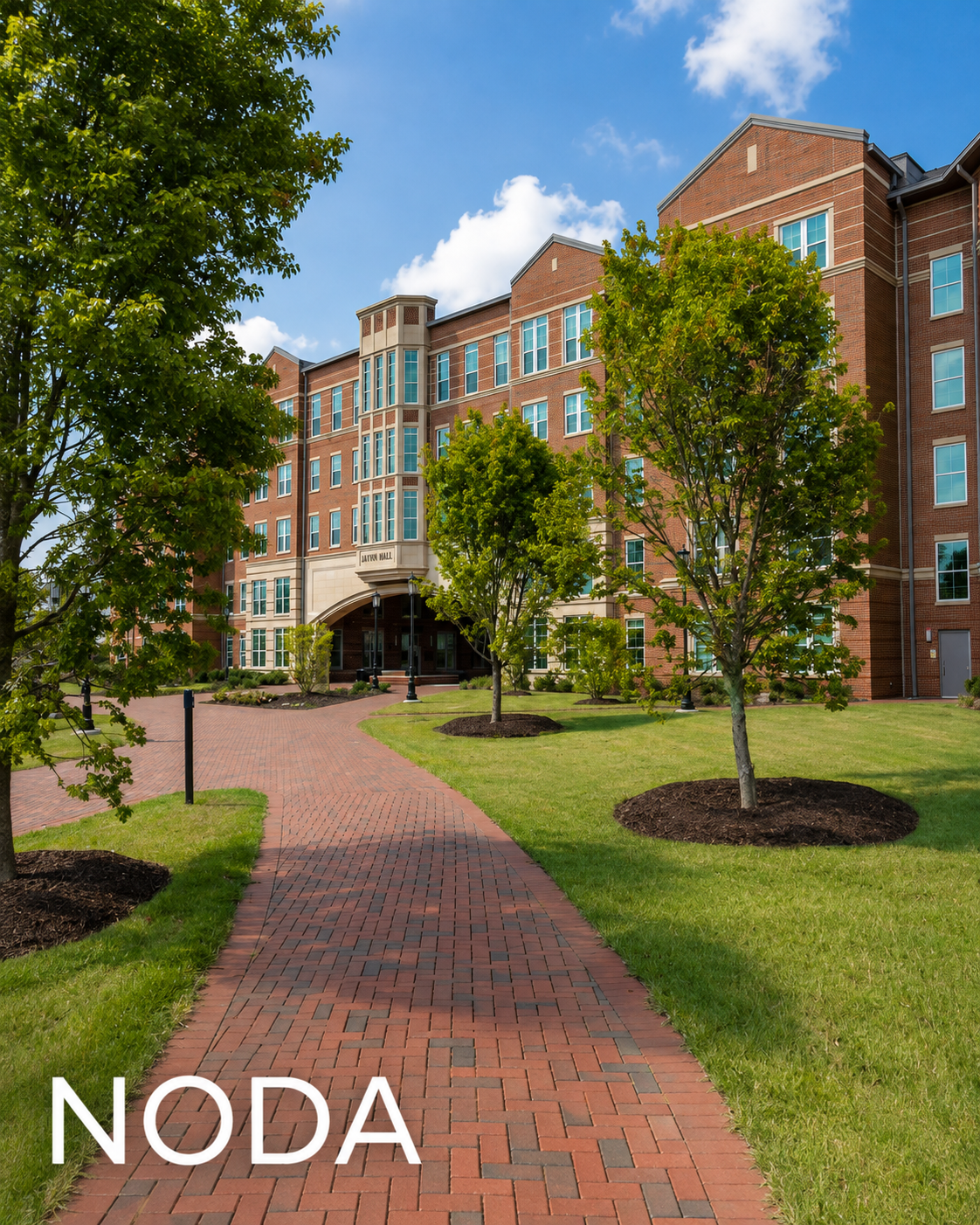 Red brick apartment building with green trees and a brick walkway in front, under a partly cloudy blue sky.