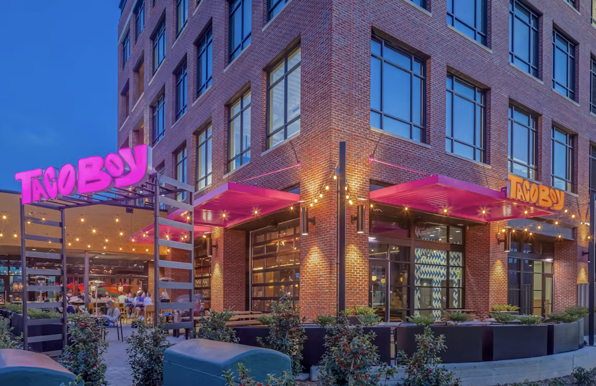 Exterior view of a modern brick building with illuminated pink and orange signage that says 'Taco Boy,' outdoor seating, string lights, and a clear blue sky in the evening.