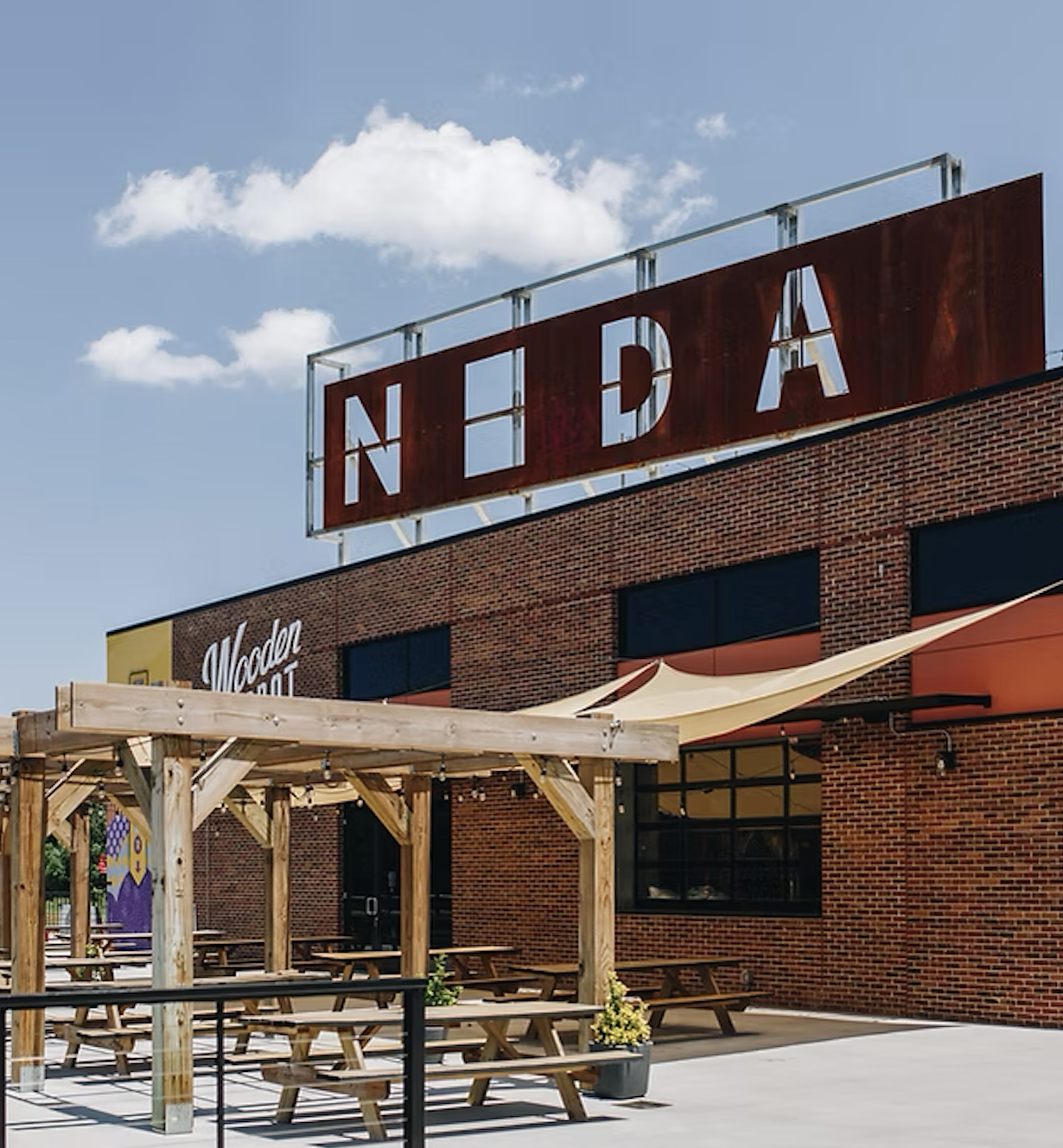 The exterior of a restaurant building with a large, weathered red sign that says "NEDA." In front, there is a wooden pergola with picnic tables underneath and a yellow awning attached to the building.