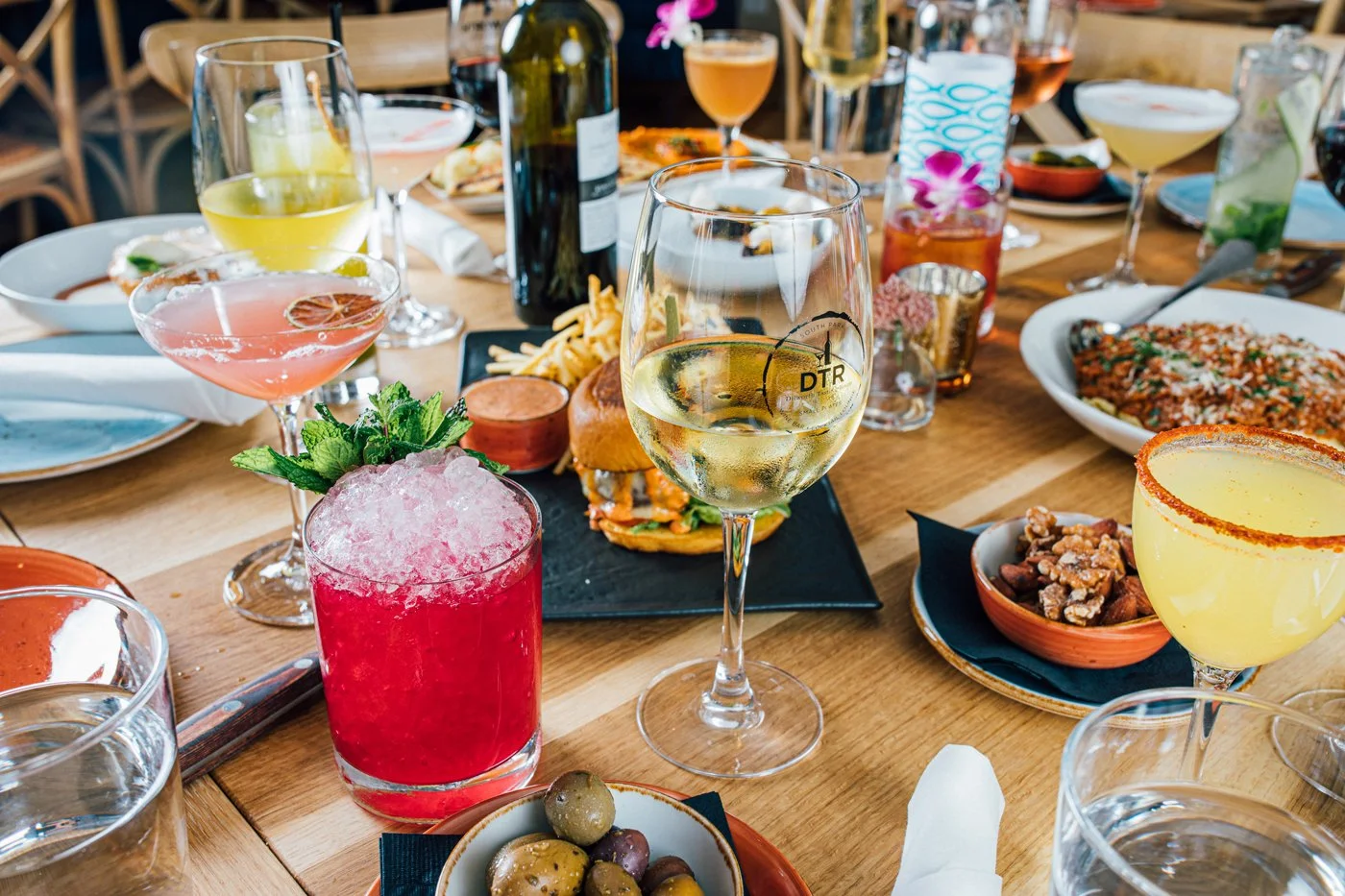 Dining table with various cocktails, wine, food dishes, and appetizers at a social gathering.
