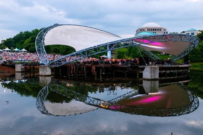 Modern outdoor concert pavilion with a white and pink-lit canopy, reflected in a calm body of water, with a crowd in the background.