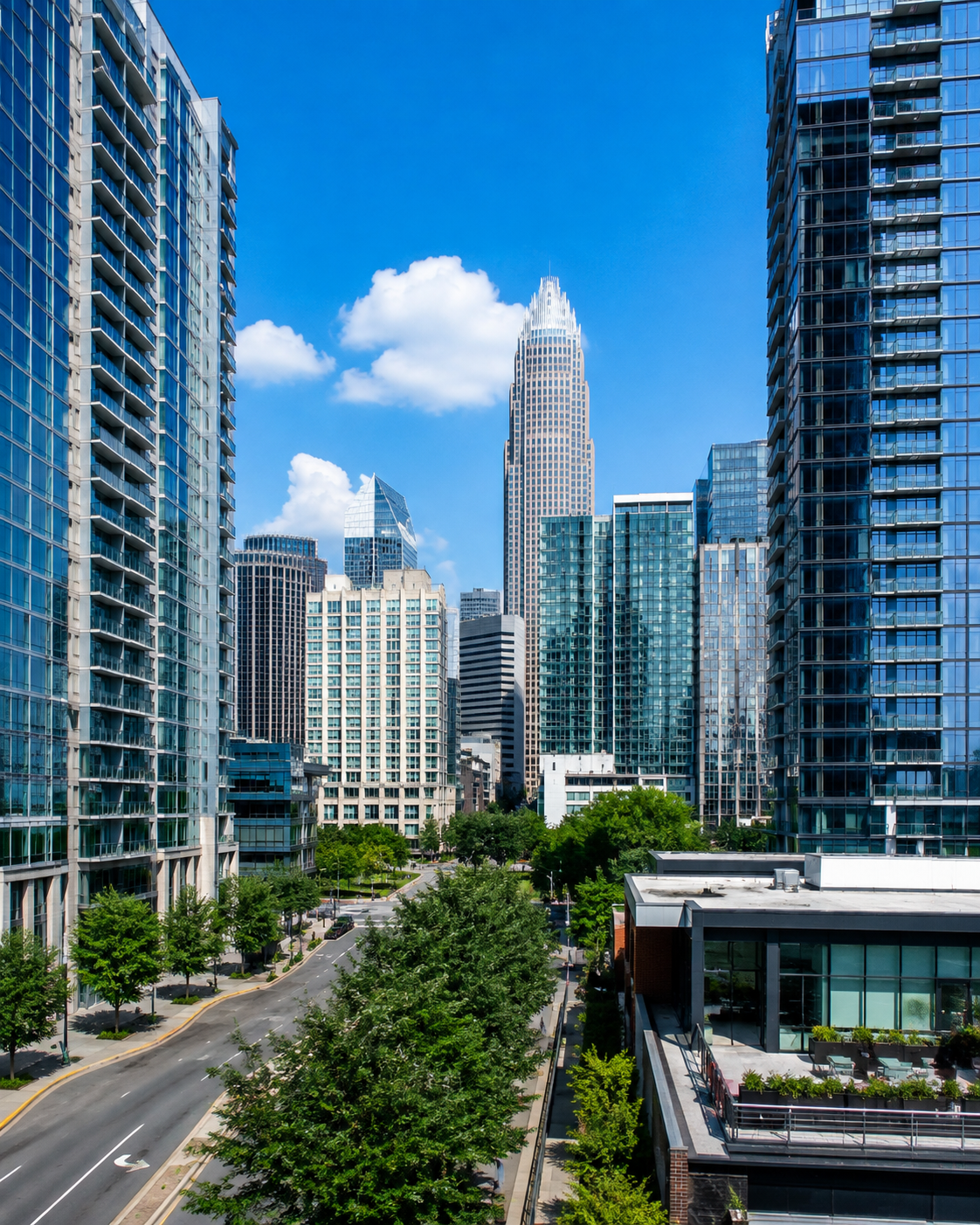 Cityscape with tall modern glass buildings, a blue sky with some white clouds, and green trees along the streets.