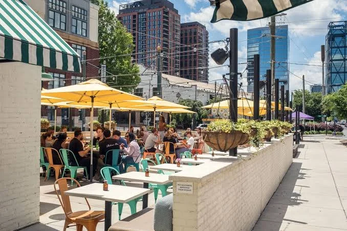 Outdoor sidewalk cafe with people dining under yellow umbrellas in an urban setting, with modern high-rise buildings in the background.