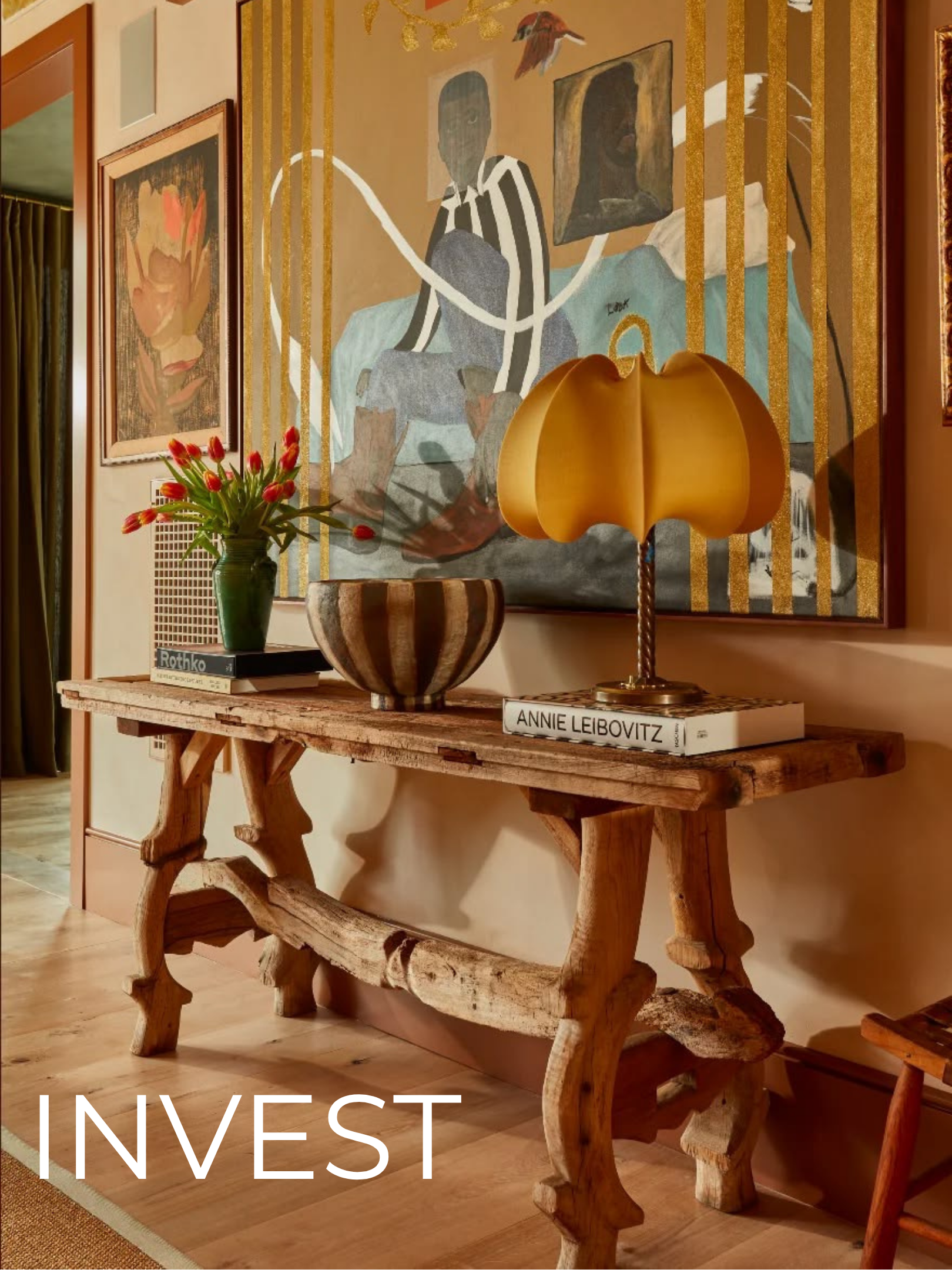 A wooden console table with curved legs in a room, decorated with a green vase holding red tulips, a large wooden bowl, and a white art book titled 'Annie Leibovitz.' Above the table is a large abstract portrait painting of a person with dark skin, wearing striped clothing, sitting in a room with framed artwork and a lamp in the background.
