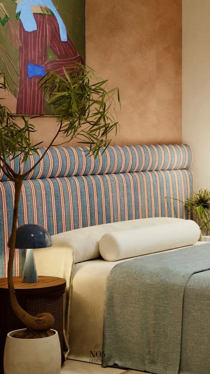 Interior of a living room or sitting area with a striped upholstered headboard, a beige sofa with a cylindrical pillow, a small side table with a blue lamp, a potted plant, and a colorful painting on the wall.