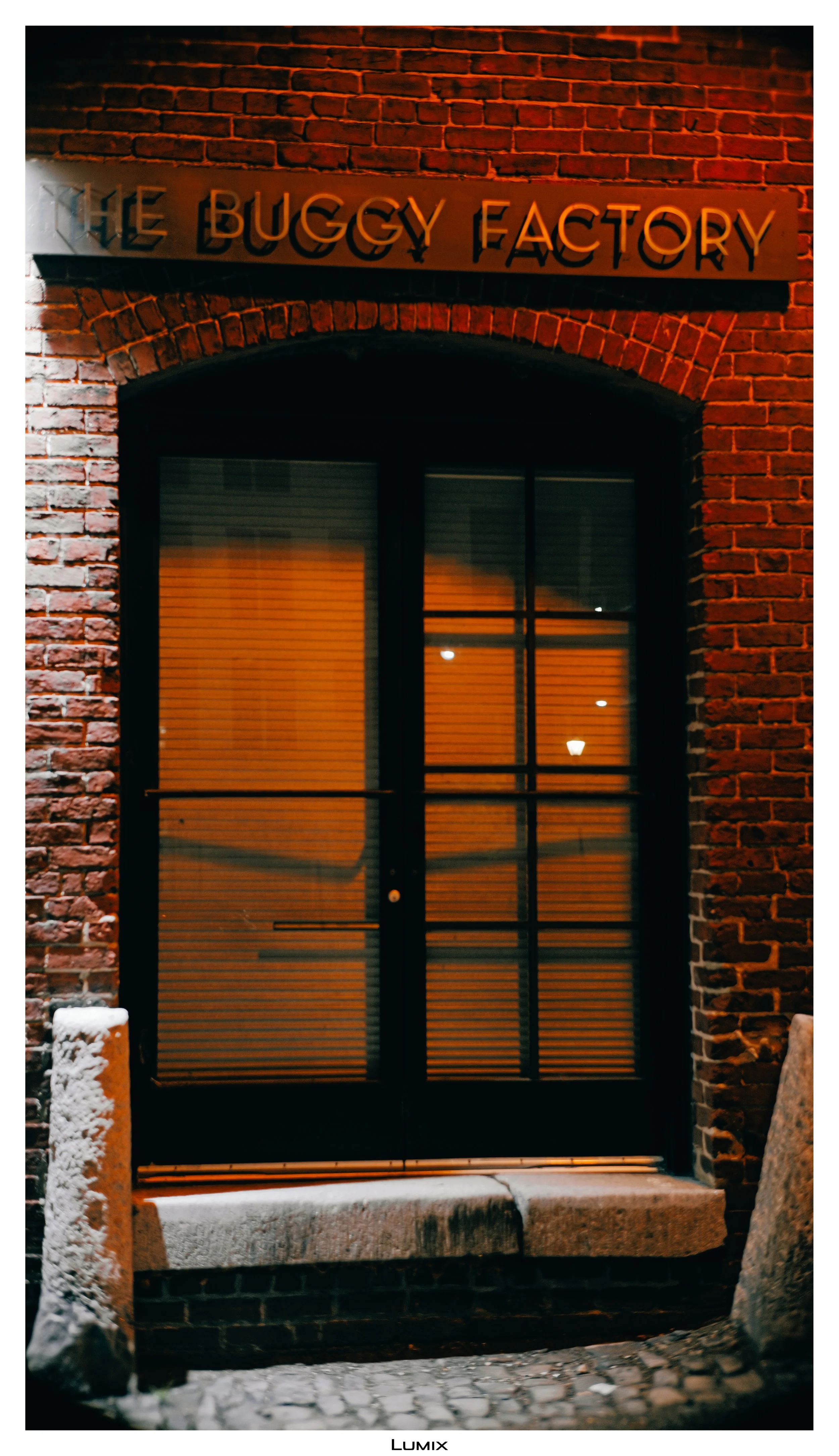 Night view of a brick building entrance with a glass door and a sign that reads 'The Buggy Factory' above the door, illuminated by warm lighting.