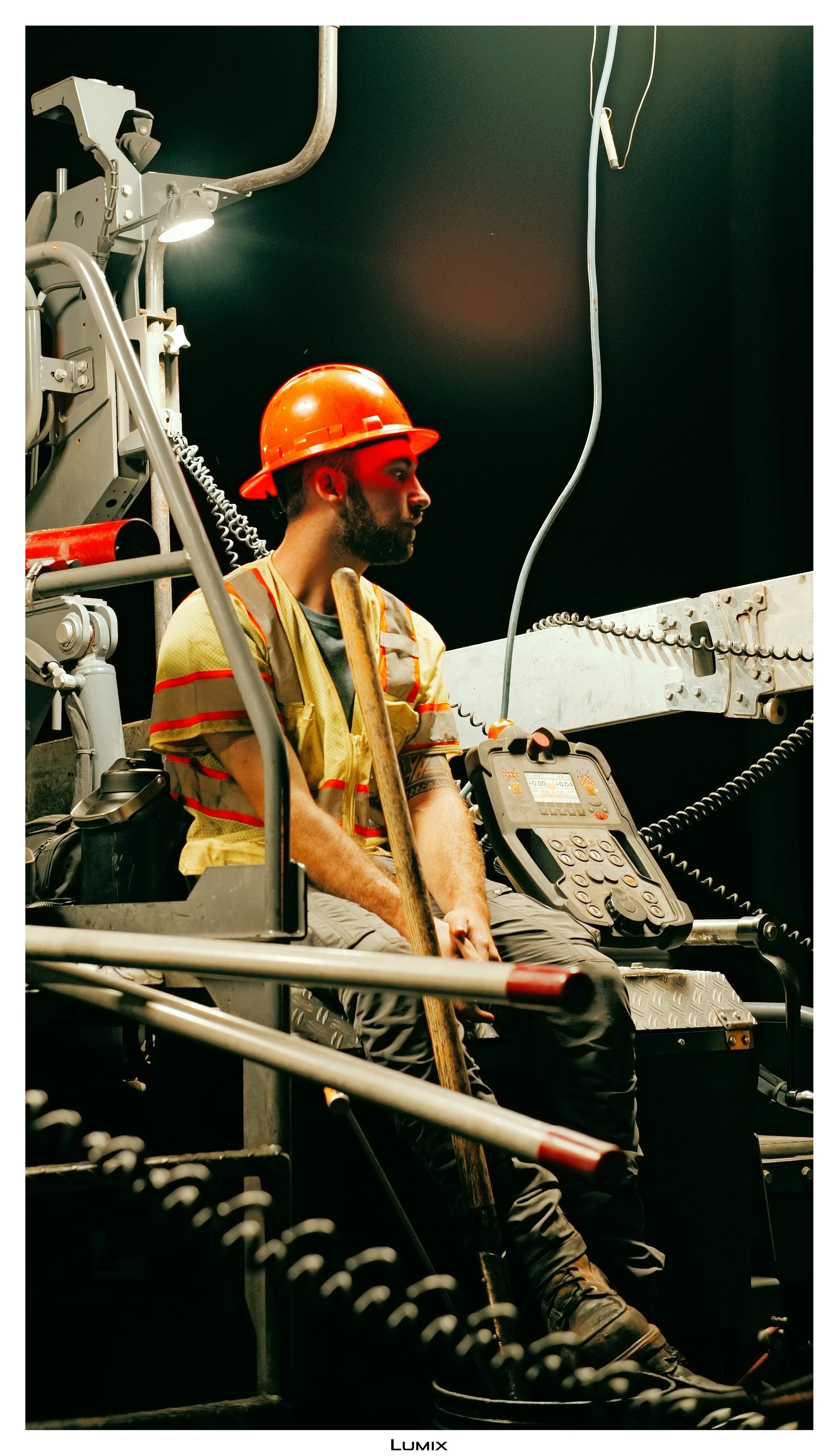 A man in safety gear, including an orange hard hat and yellow vest, sitting on industrial equipment in a control room or workshop, with various machinery, cables, and a control panel around him.