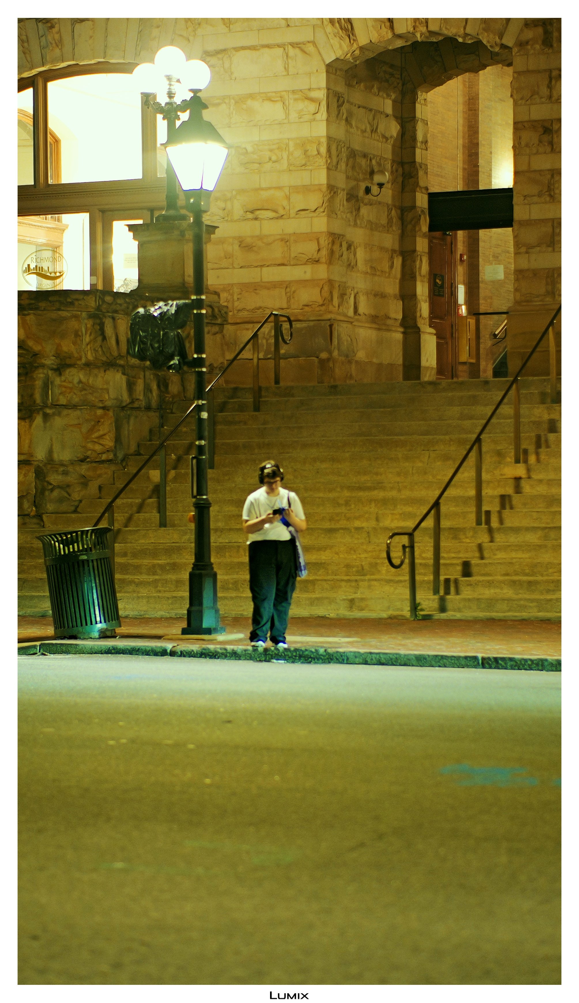 A person in white shirt and dark pants standing on sidewalk at night, holding a handheld device, with stone staircase, lamp post, and building in the background.