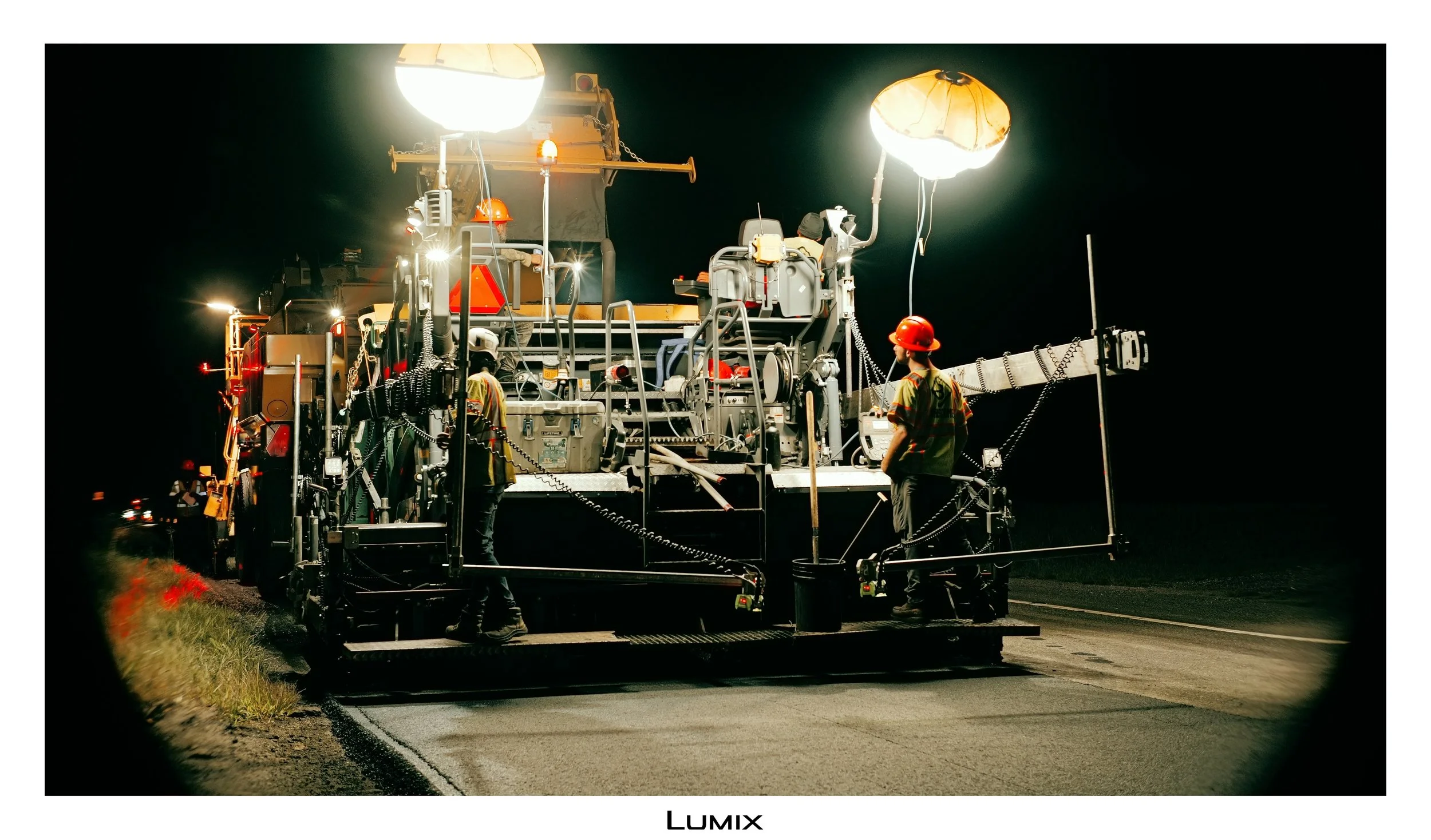 Night scene of road work with large illuminated equipment and workers in safety gear paving or repairing the road.