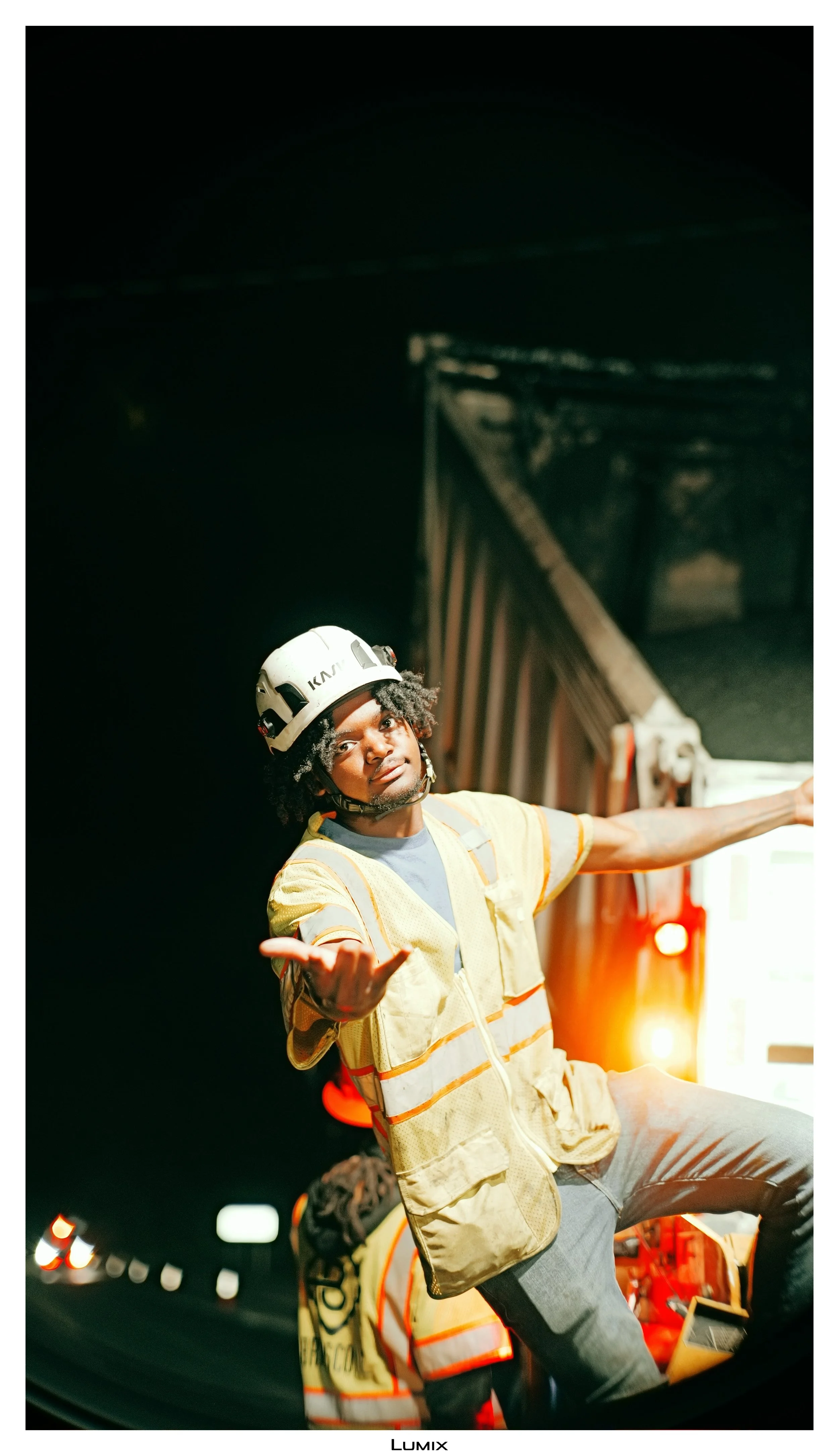 A young male construction worker in a yellow safety vest and a white hard hat is gesturing with his hands while standing on a street at night. He is illuminated by bright lights, and a large vehicle, possibly a fire truck, is in the background with i