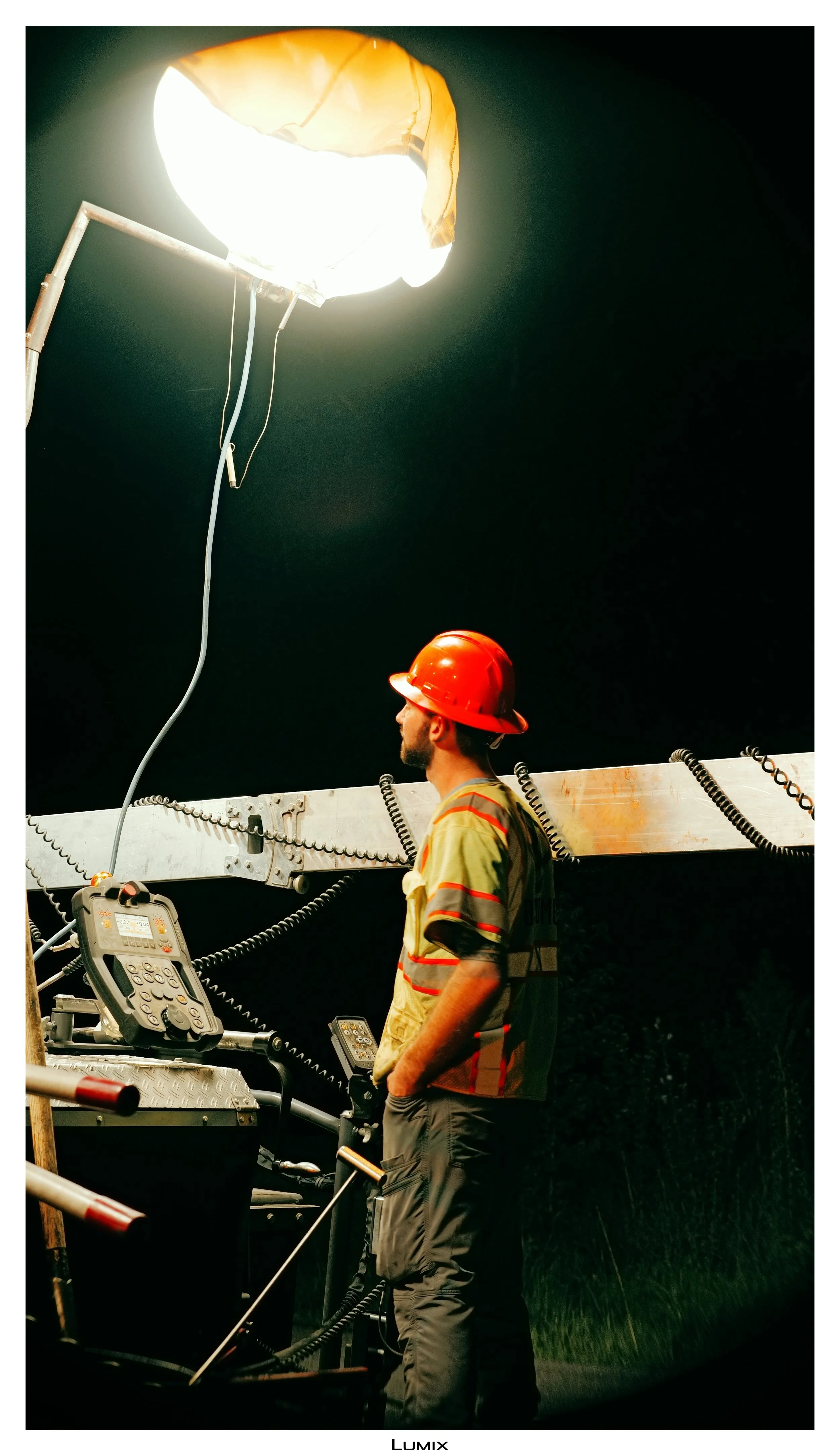 A construction worker wearing a red safety helmet and yellow shirt with reflective stripes is standing next to equipment with communication devices. Above him, a large, bright light is shining downward, illuminating his side profile against a dark ba