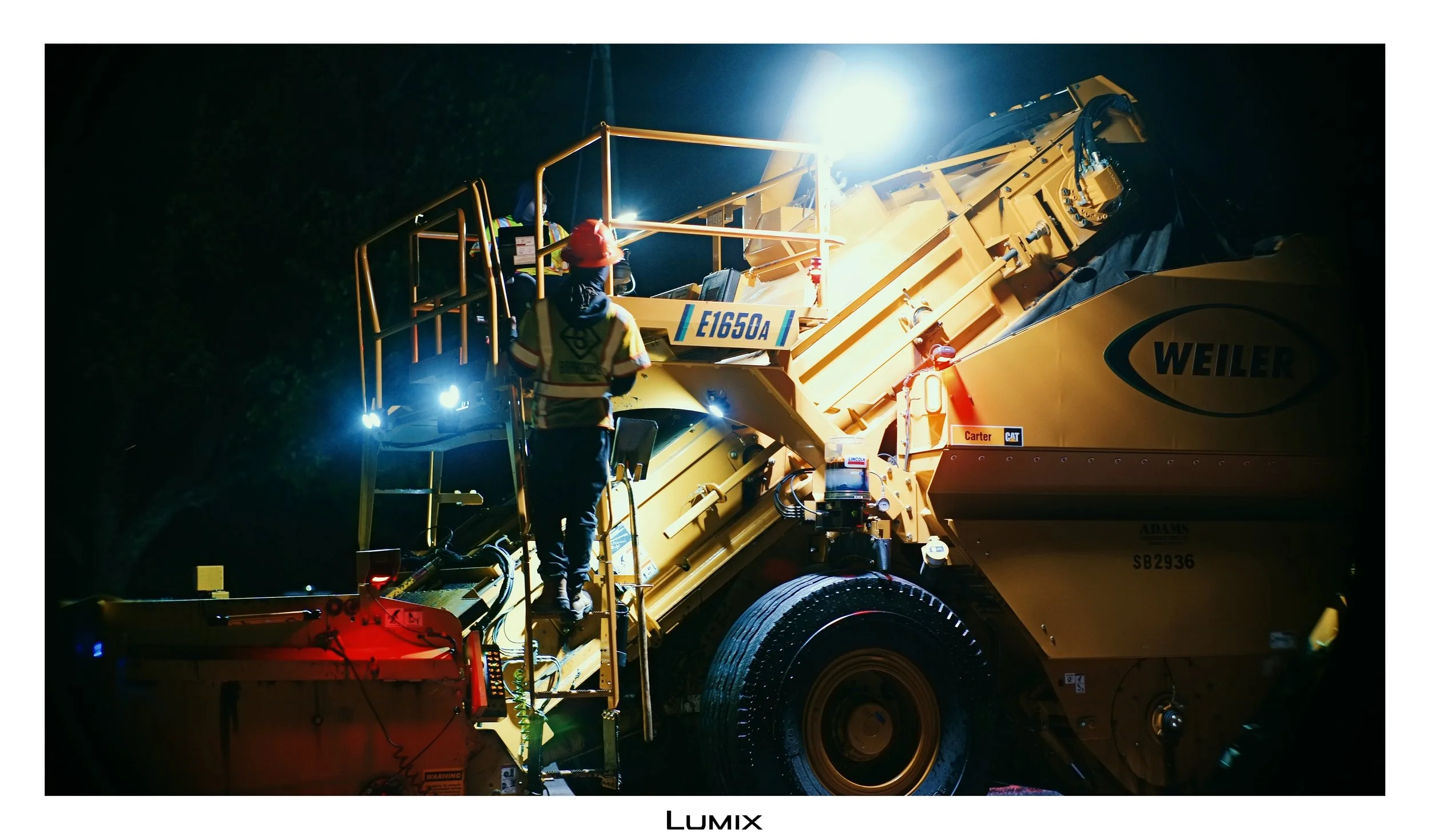 Nighttime scene showing construction workers operating heavy machinery, specifically a large yellow Caterpillar machine, illuminated by bright lights, with workers wearing safety vests and helmets.