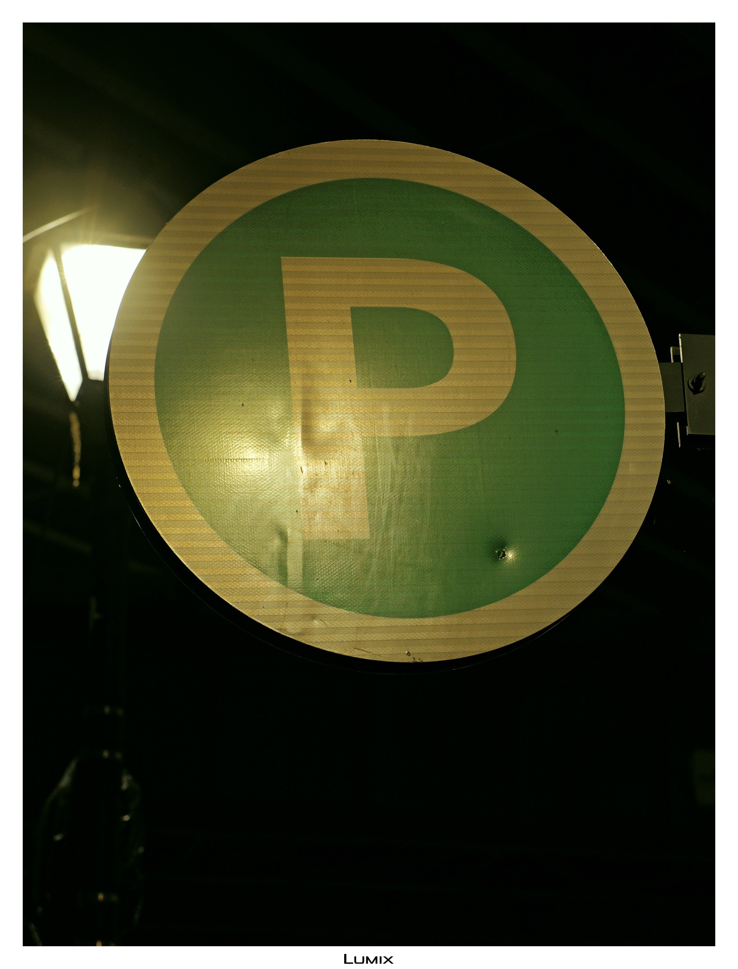 Close-up of a round green and yellow sign with a letter 'P' in the center, illuminated by a bright light source from the left side, mounted on a dark background.