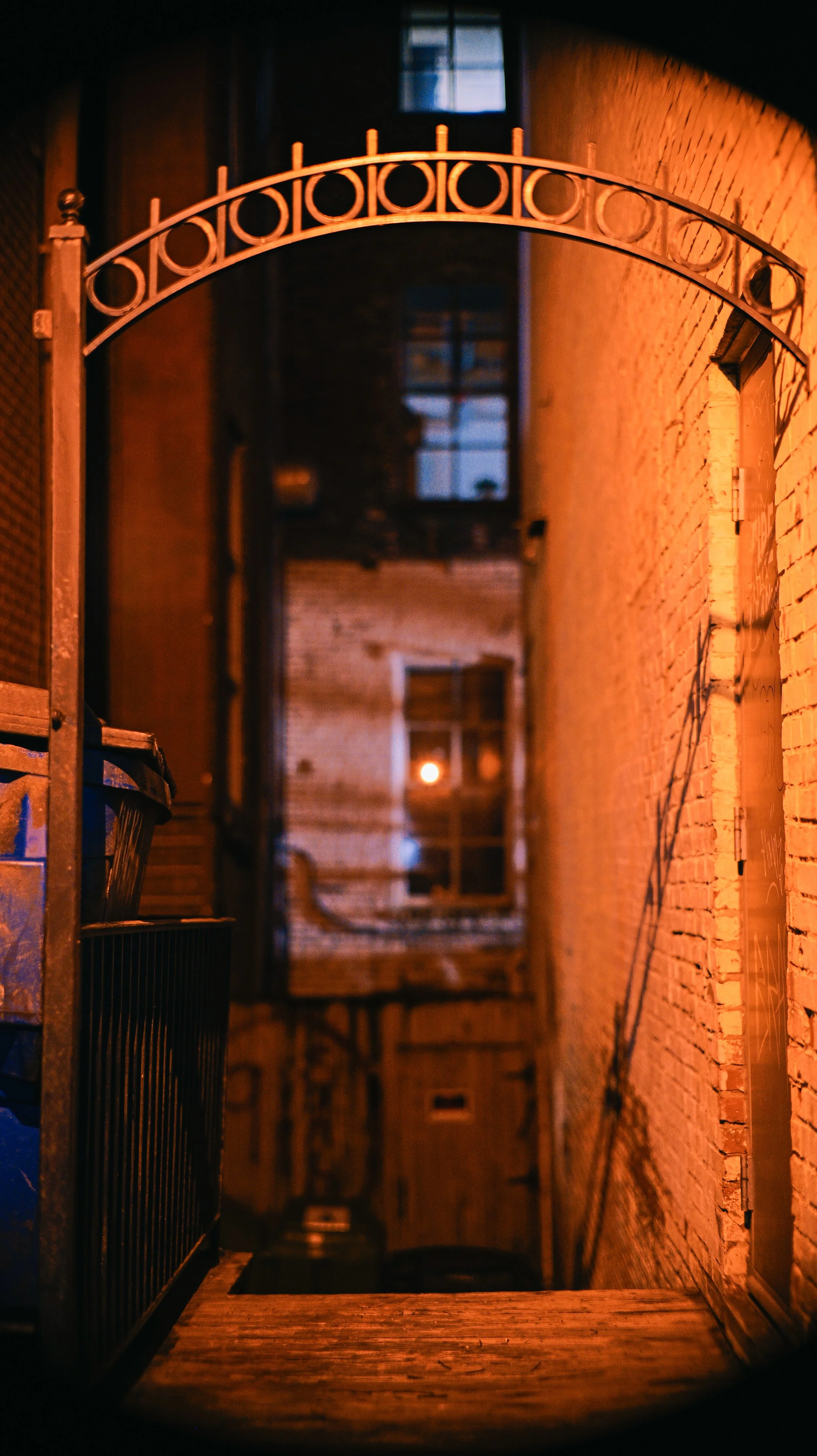 Nighttime alleyway with brick walls, an arched metal gate, and a lit window in the background.
