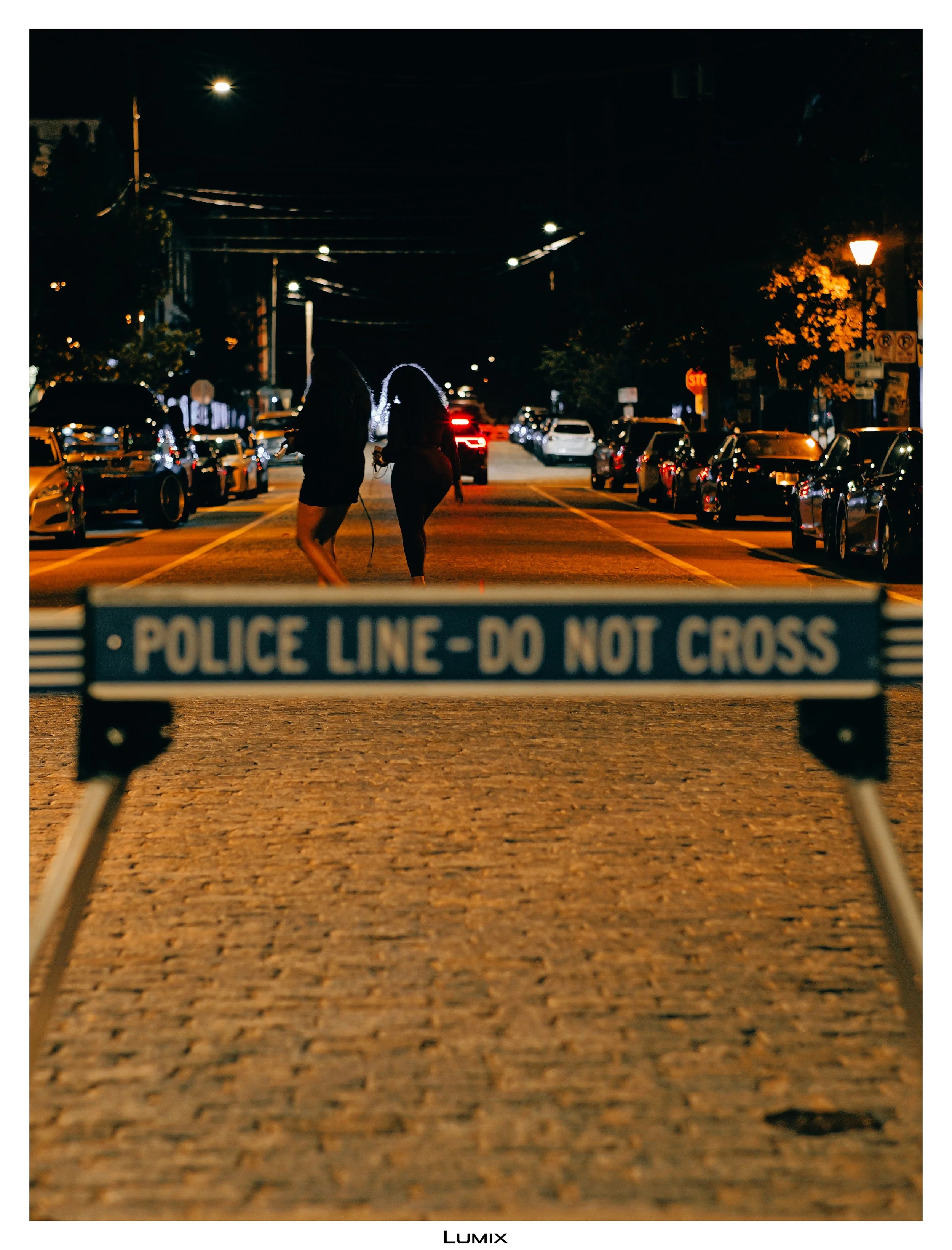 Night scene on a city street with cars parked along both sides, streetlights illuminating the area, and two people walking near a police line caution tape in the foreground. The street appears quiet and dark overall.