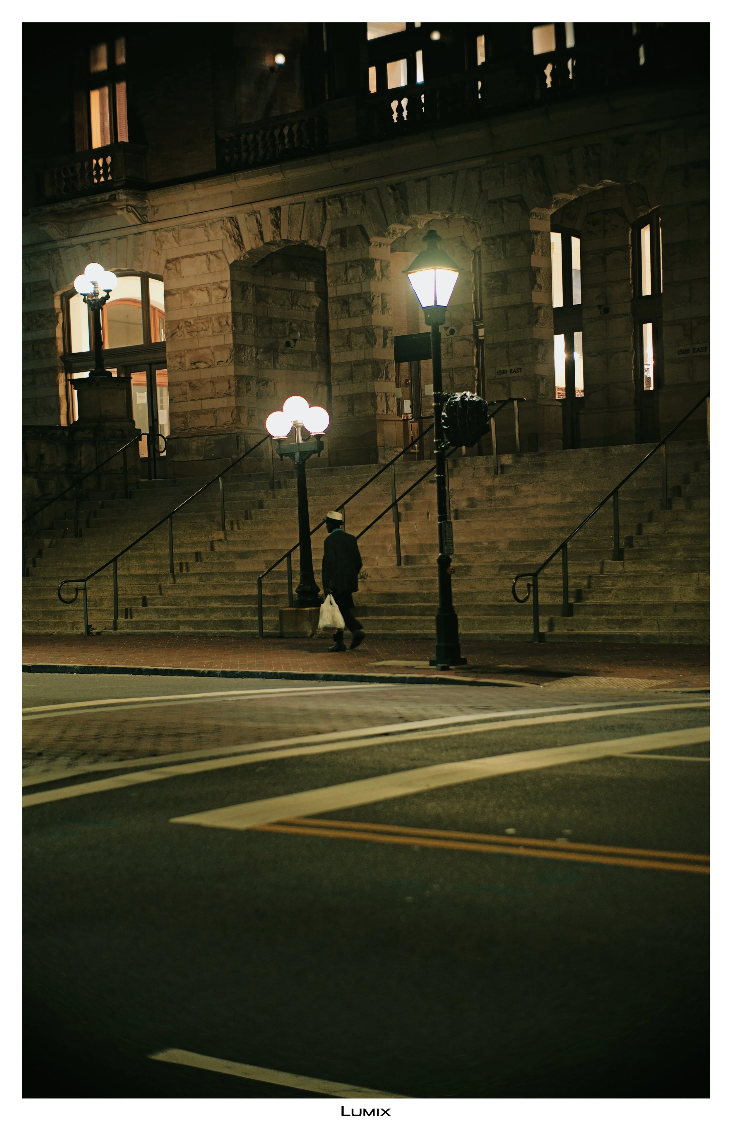 Nighttime scene of a stone building with steps leading up to its entrance, lit by street lamps. A person wearing a hat, carrying a bag, walks on the sidewalk.