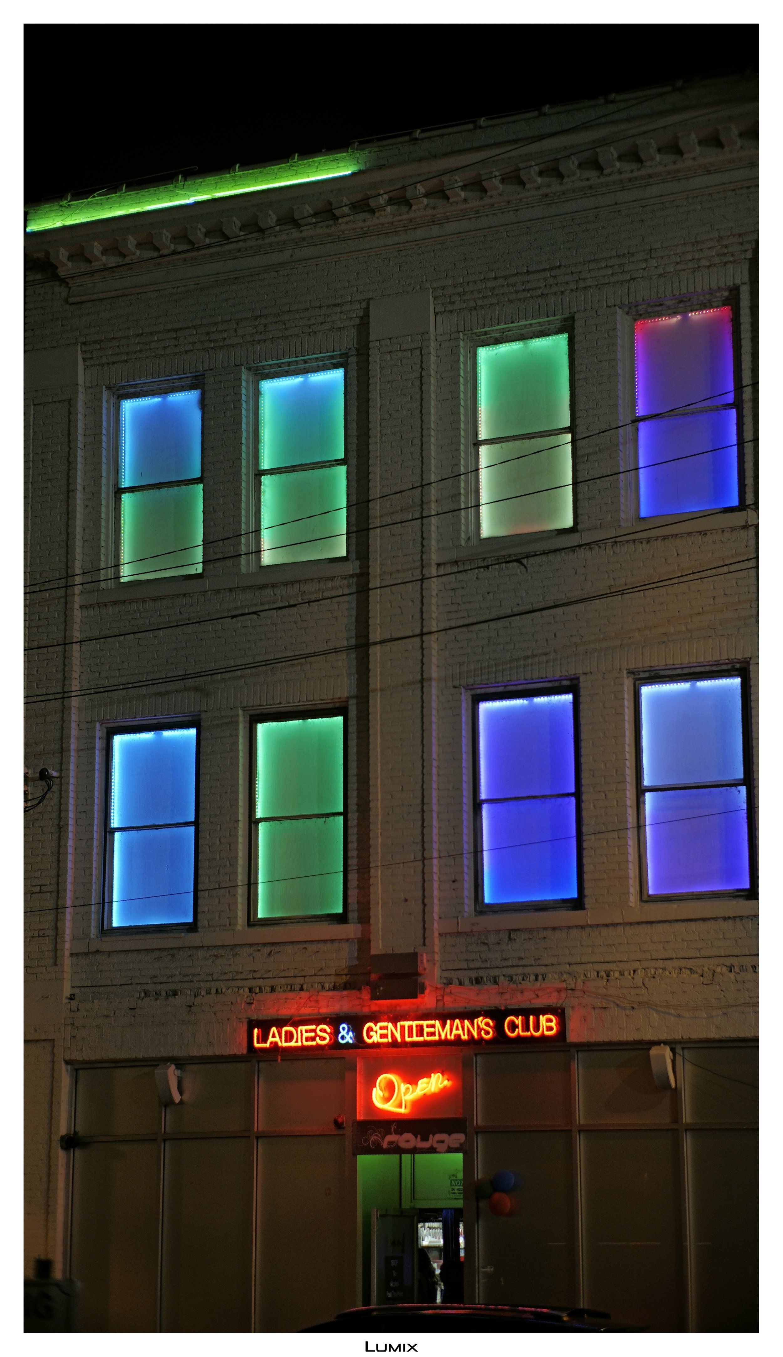 The building has a row of five windows illuminated with colorful neon lights: blue, cyan, green, purple, and blue. A neon sign reads 'Ladies & Gentleman's Club' with an additional 'Open' sign beneath it.