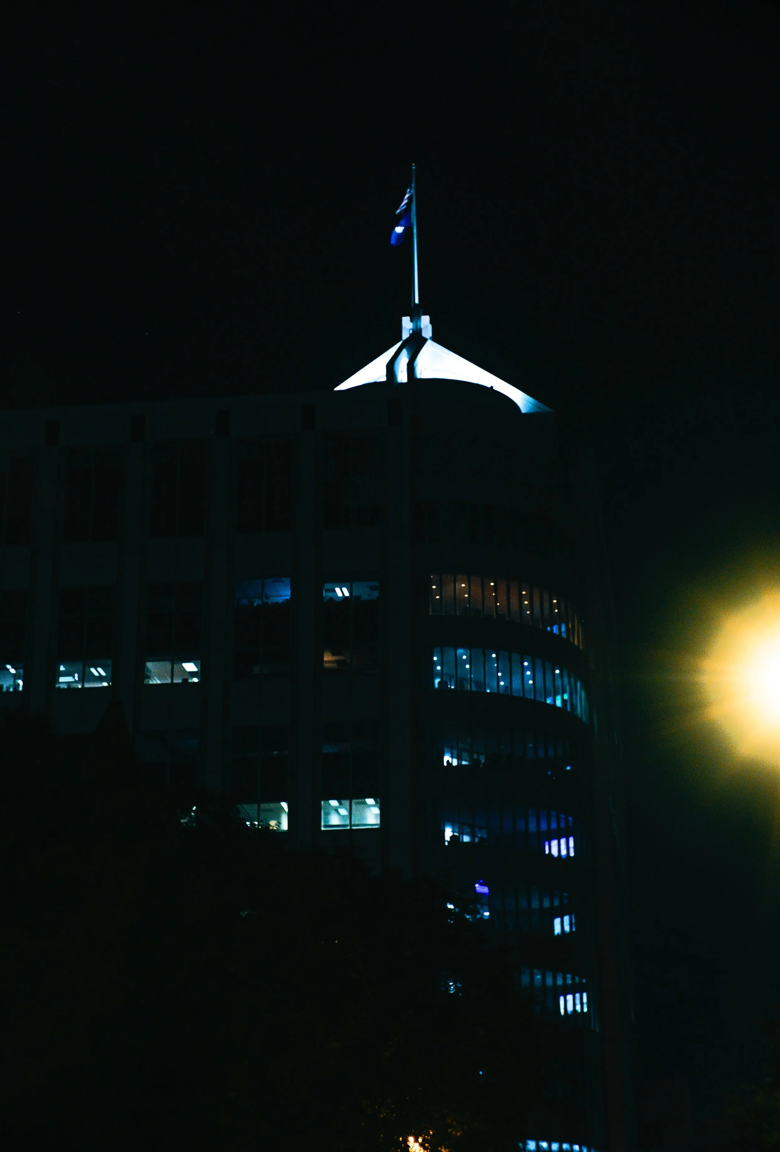 Nighttime photo of the top of the White House with an illuminated flagpole and the American flag waving, against a dark sky with some stars visible.