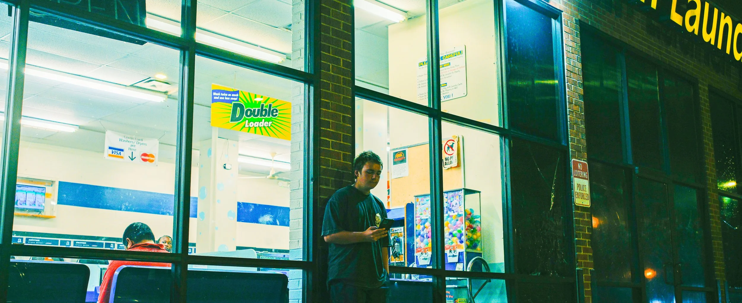 A young man standing outside a laundromat at night, looking at his phone, with a bright interior visible through large glass windows, including laundry machines, and signs advertising laundry services.