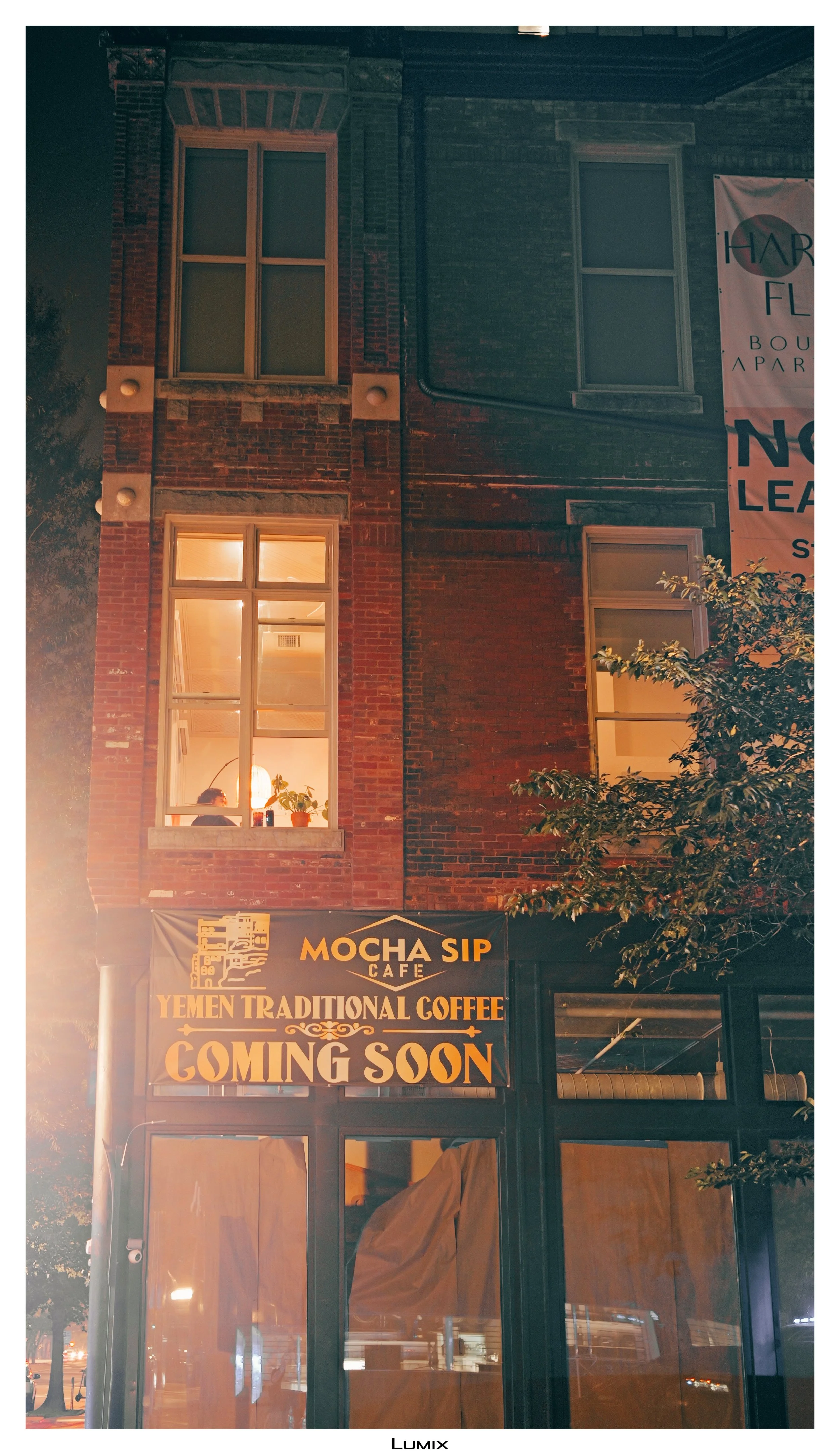 Nighttime view of a brick building with illuminated windows. A sign on the building reads "Mocha Sip Cafe, Yemen Traditional Coffee Coming Soon."