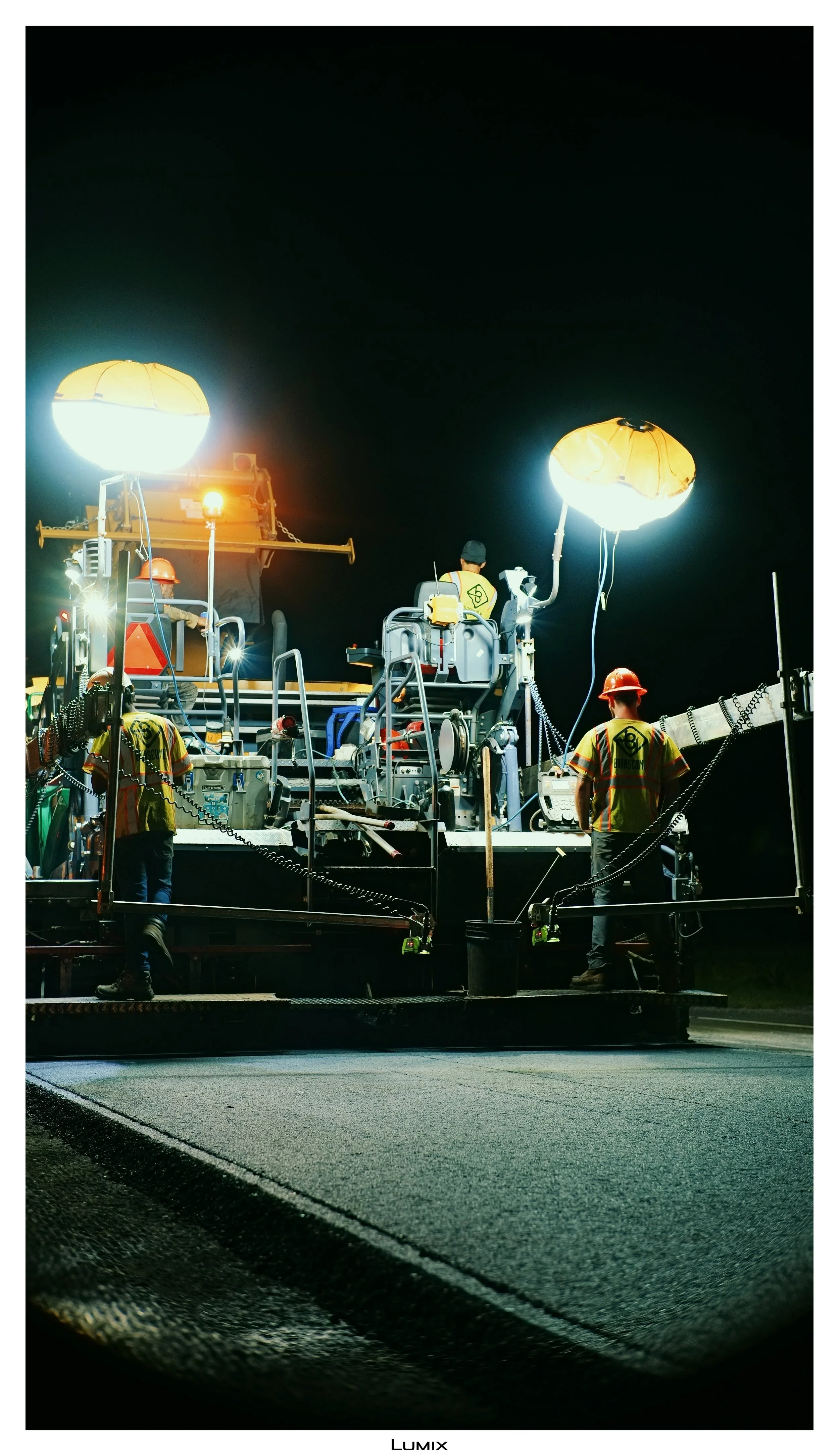 Nighttime road construction scene with workers in safety gear operating large equipment illuminated by bright work lights.