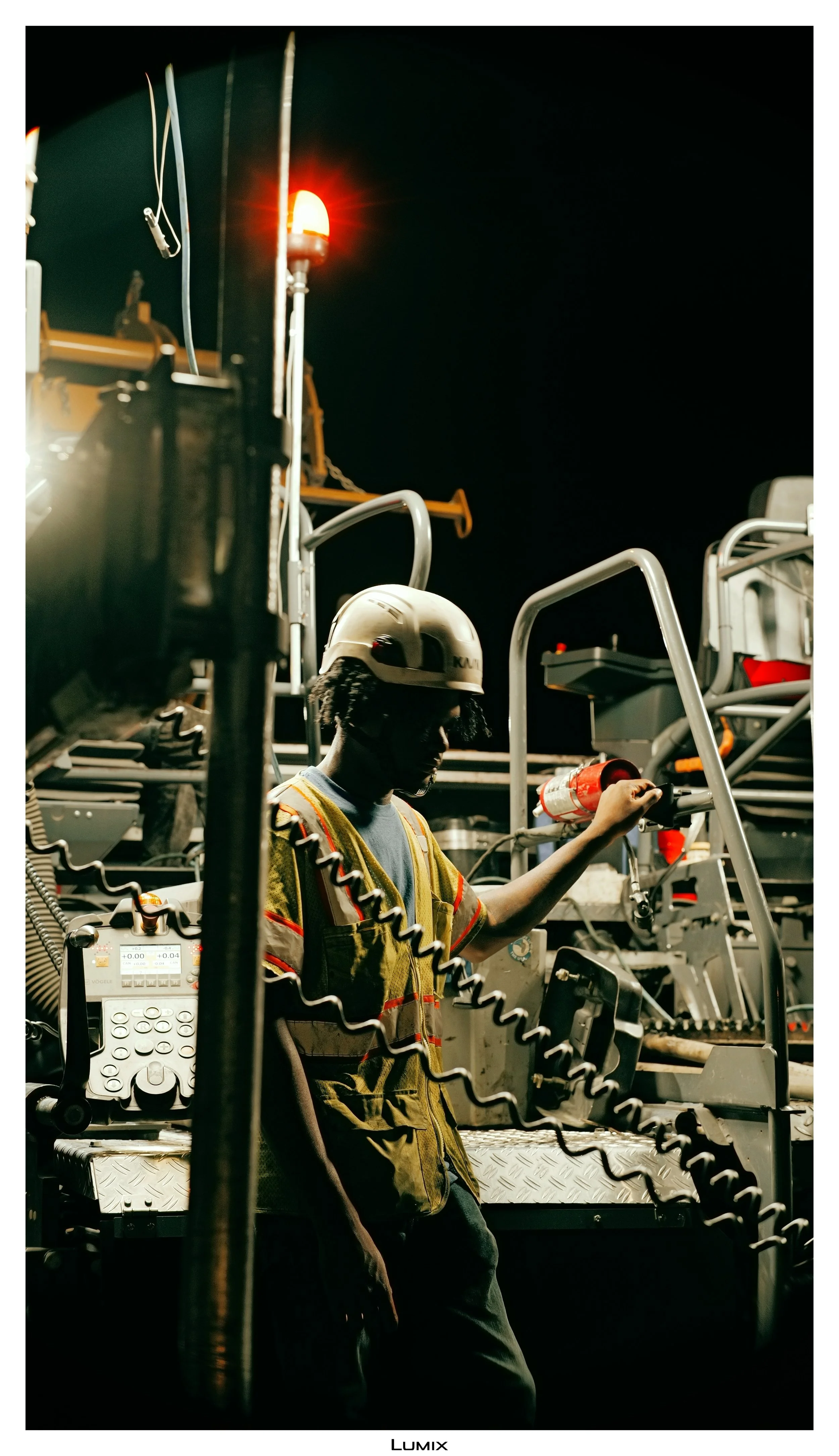 A worker wearing a helmet and safety vest operating machinery outdoors at night, illuminated by artificial lights and a red signal light.