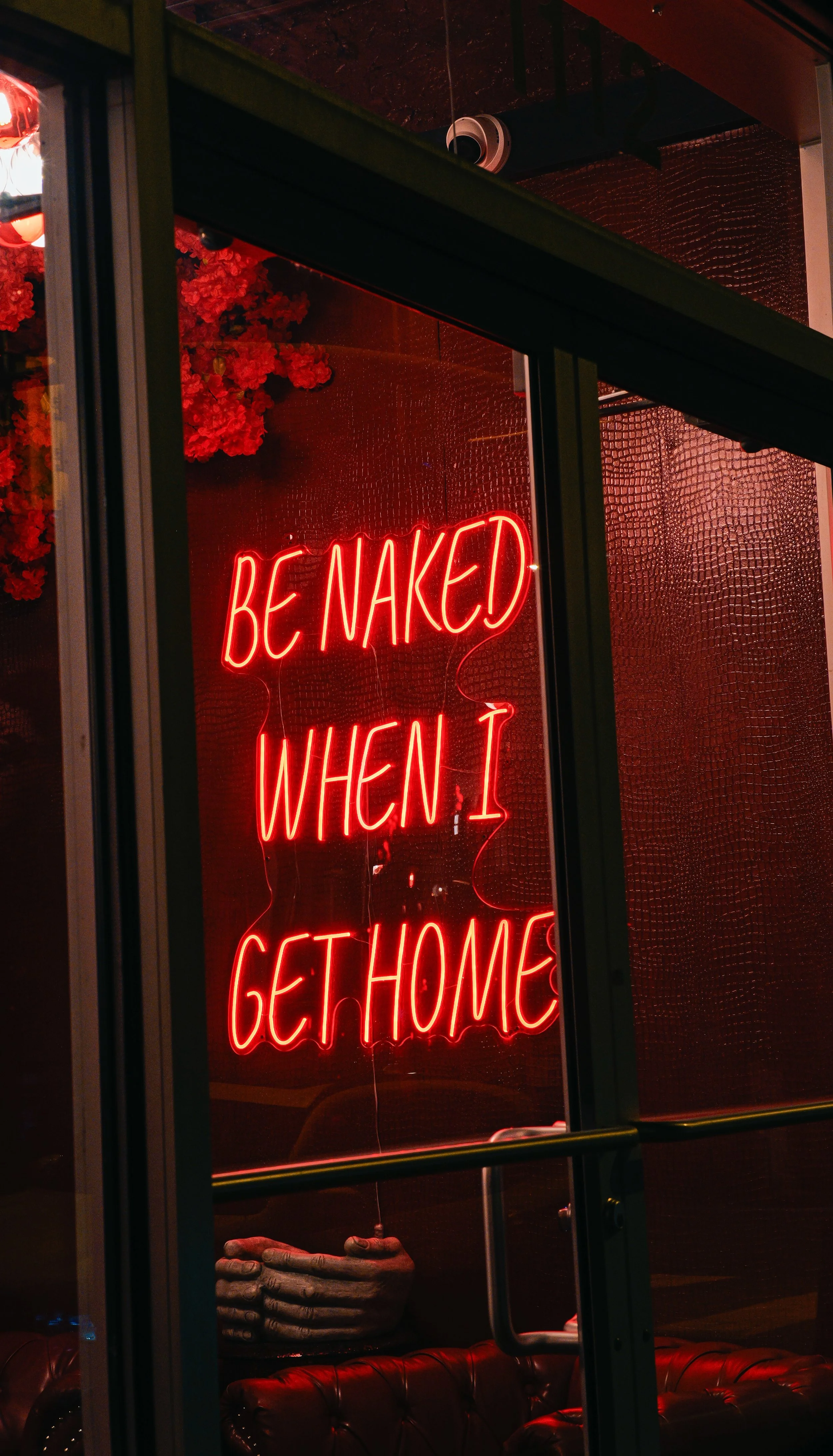 A neon sign inside a shop displays the phrase 'BE NAKED WHEN I GET HOME' in bright red. The interior has dark red walls, a leather couch, and a decorative skeleton hand resting on a table, with flowers visible in the background.