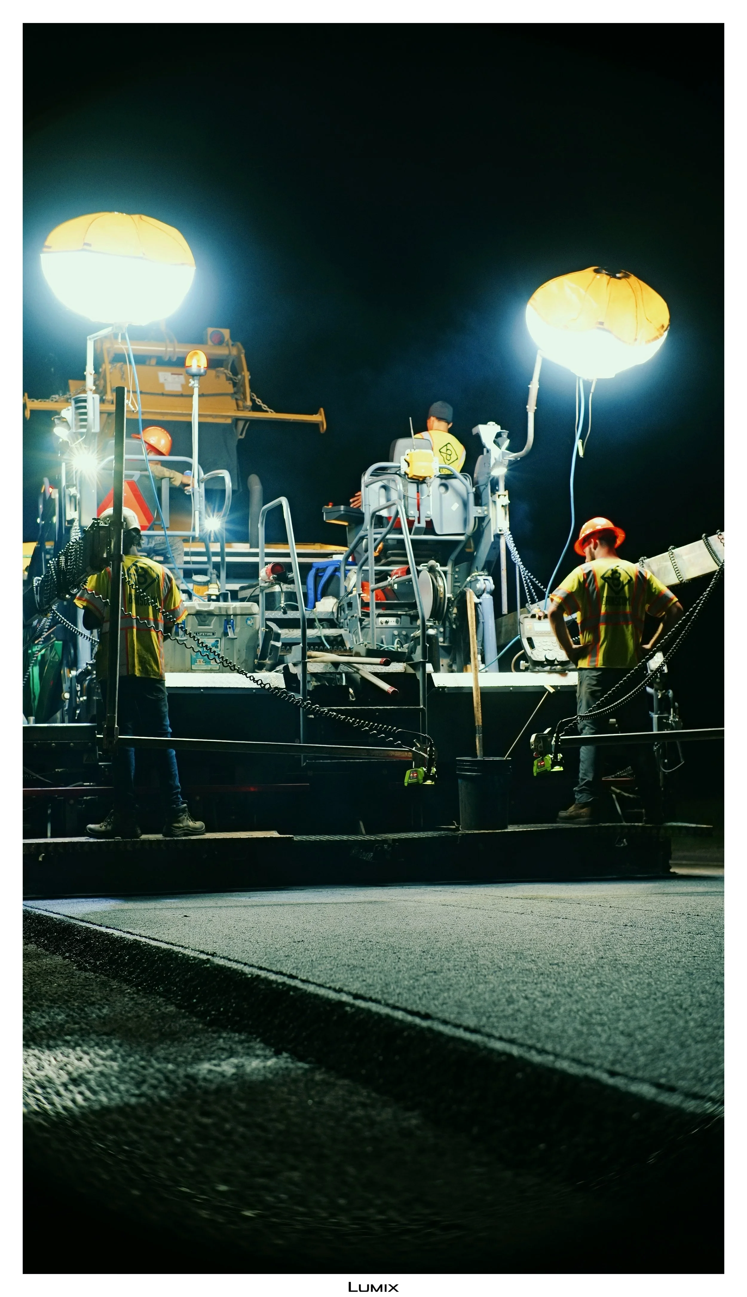 Night scene of construction workers operating equipment with bright overhead lights, working on a road or pavement.