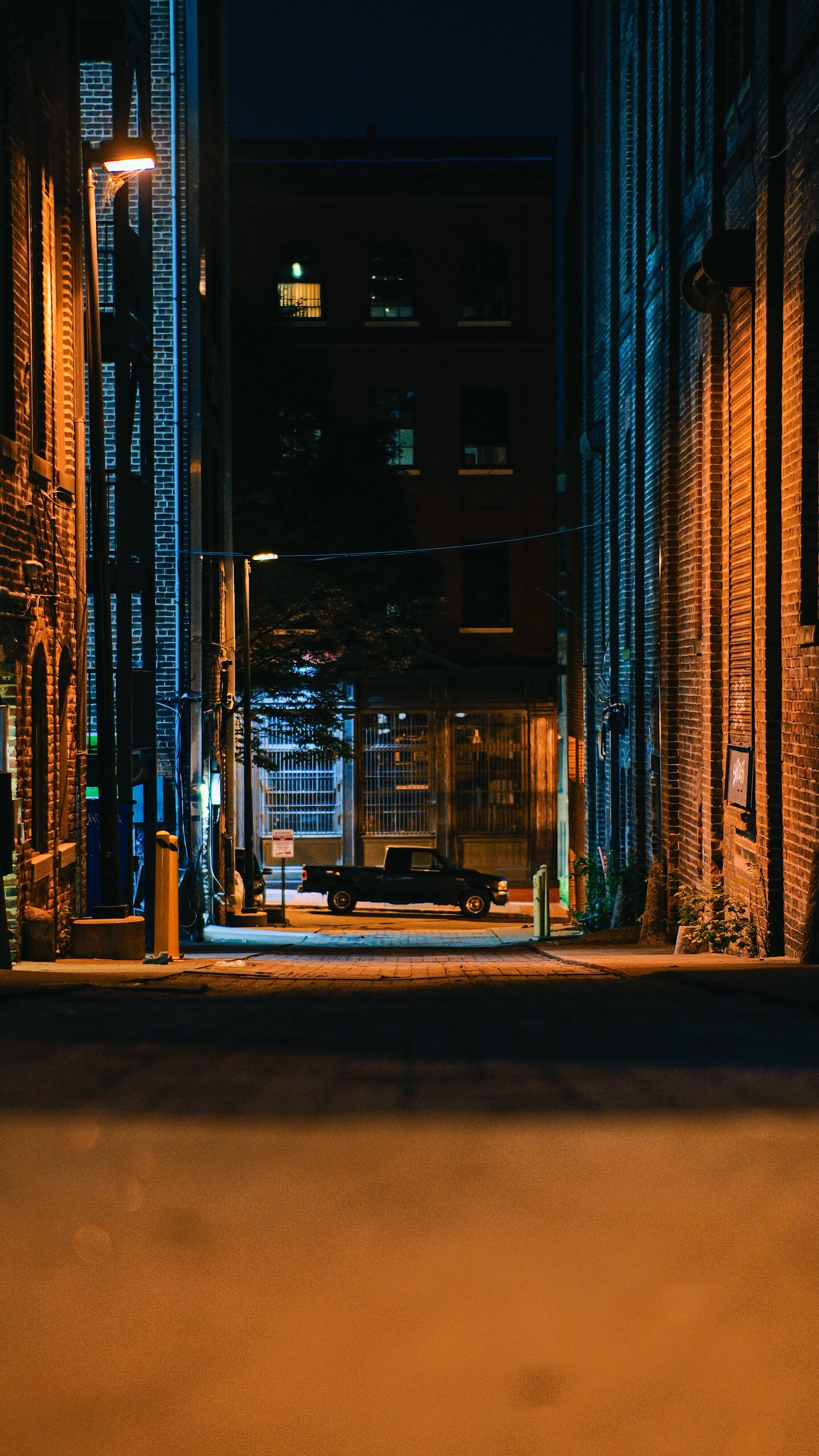Nighttime alley scene with brick buildings on both sides, a dark sky, a parked black truck, and distant lit windows.