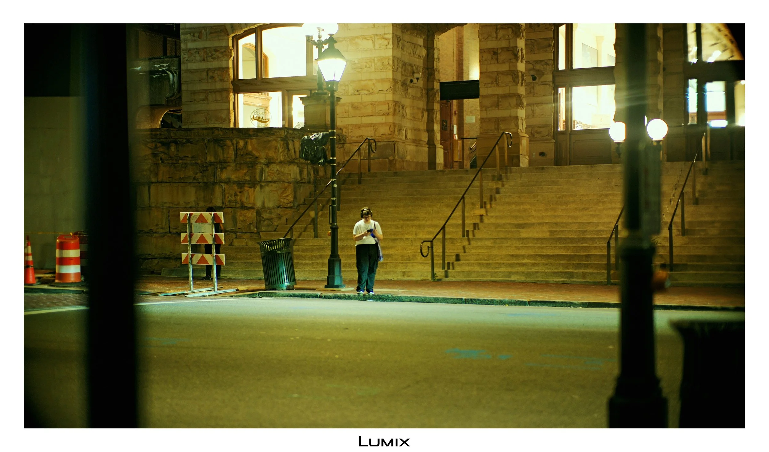 A man stands on a set of stairs outside a stone building, looking at his phone. The area is illuminated by street lamps, with construction cones and barriers nearby.