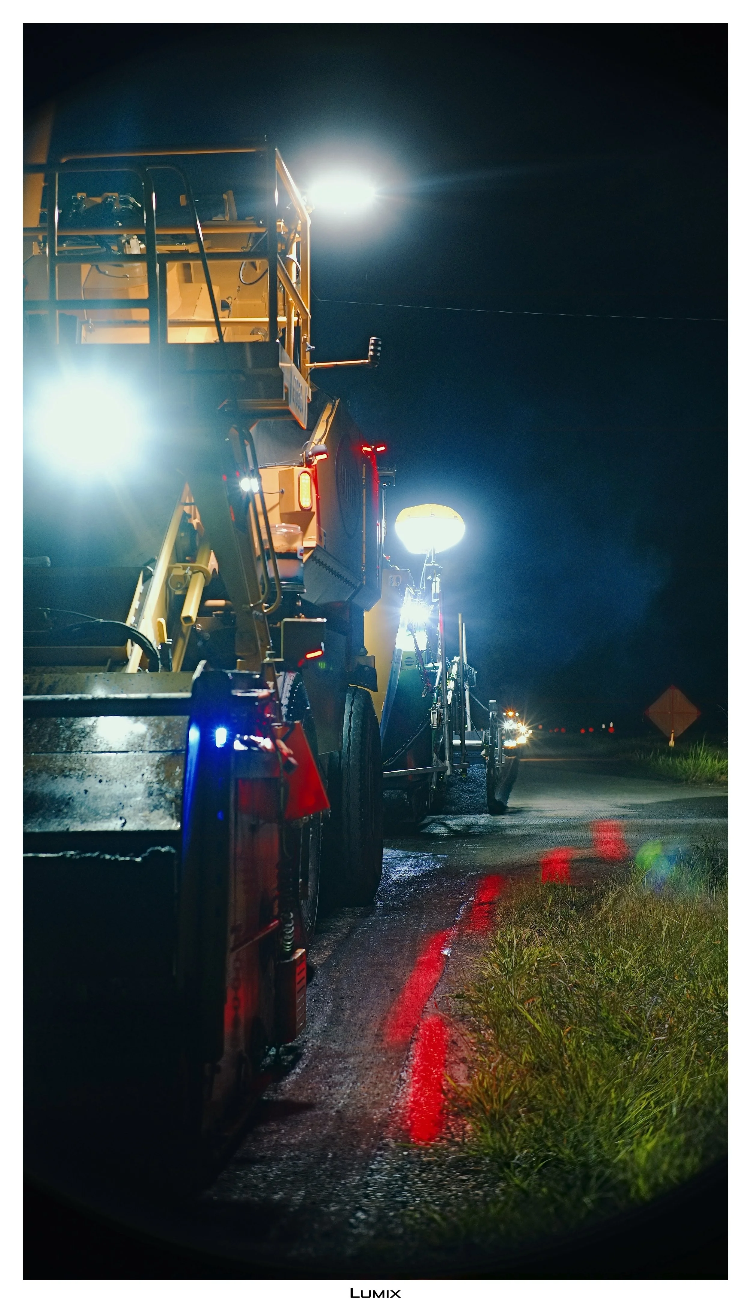 Nighttime scene with large yellow construction vehicles parked on the side of a wet road, reflecting red and white lights, with bright headlights and work lights illuminating the area under a dark sky.