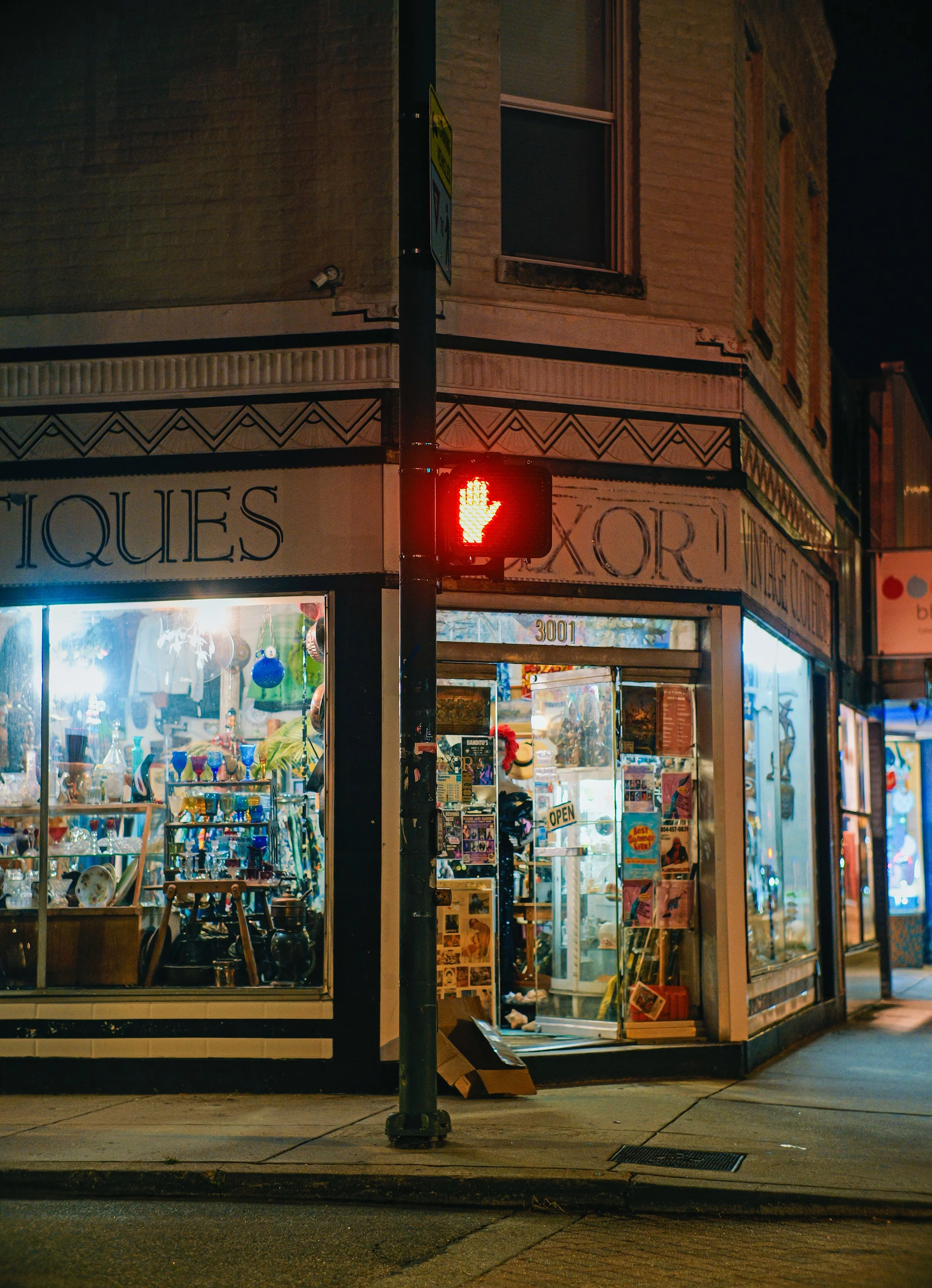 A nighttime street scene showing a corner store with large glass display windows filled with various items such as glassware, books, and decorative objects. The store has a sign with partially visible text, and the store's exterior features decorativ