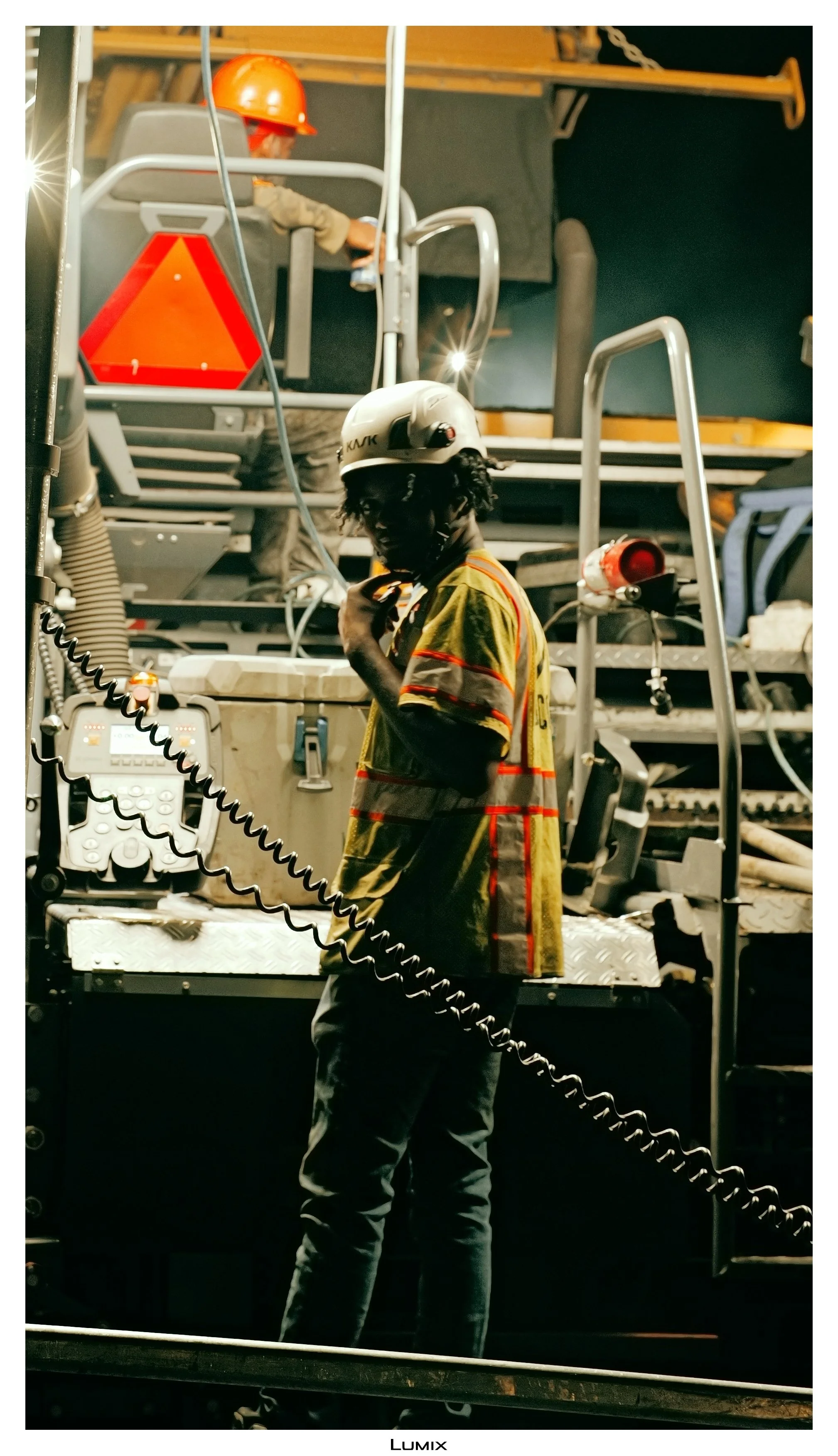 A worker wearing a yellow safety vest and helmet stands on a platform in an industrial or construction setting, with another worker in the background also wearing a helmet and operating equipment.