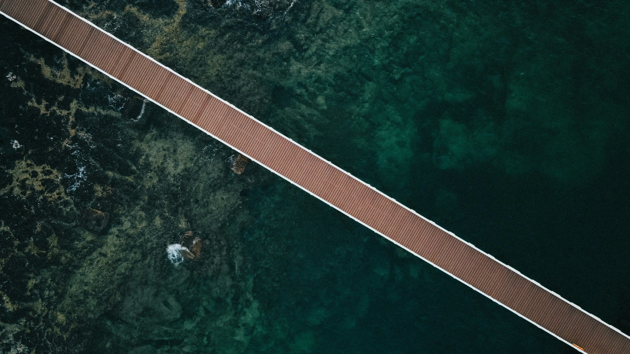 Aerial view of a wooden footbridge over clear green water and rocks.