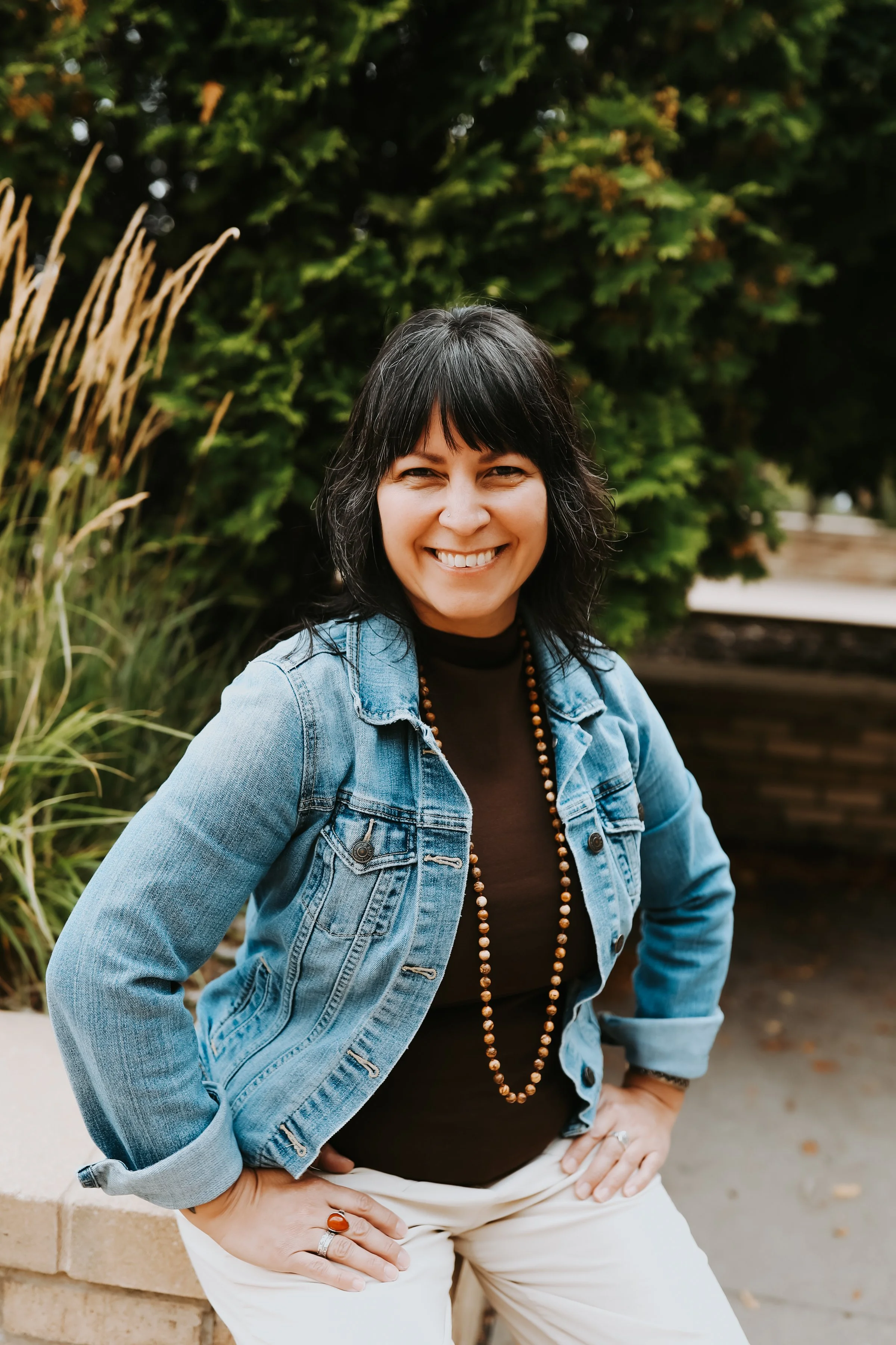 Smiling woman in a denim jacket and beaded necklace sitting outdoors in a lush garden