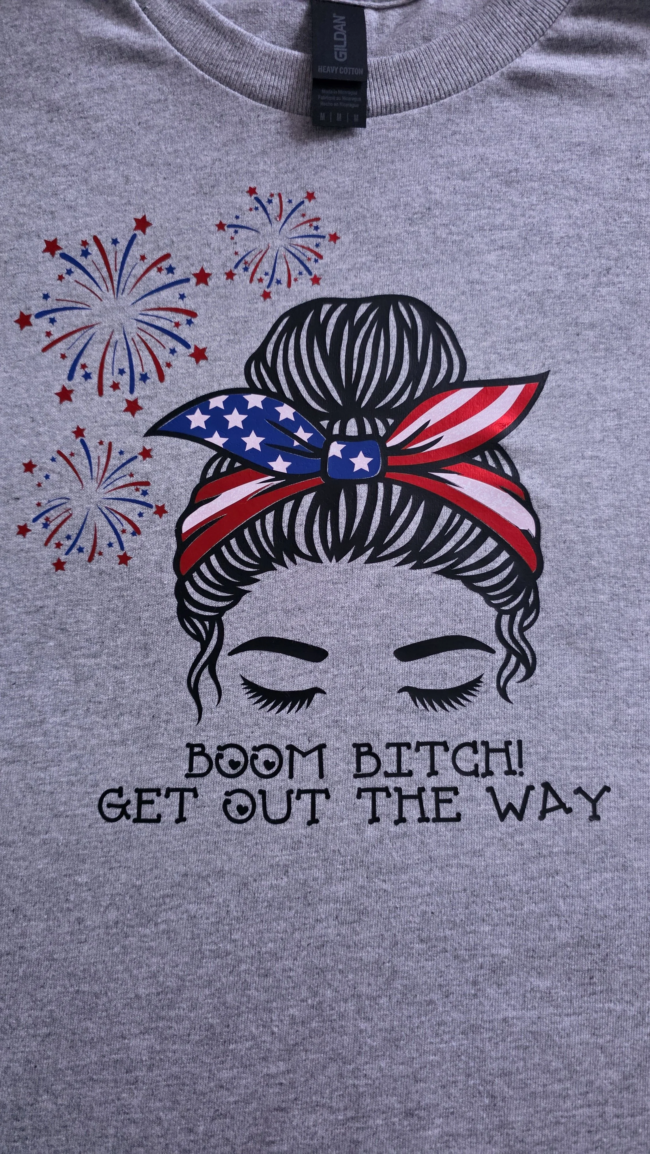 A gray T-shirt with a graphic of a woman's face with closed eyes, a messy bun hairstyle, and a bandana with an American flag pattern. Fireworks are displayed above the head. The text reads, "BOOM BITCH! GET OUT THE WAY."