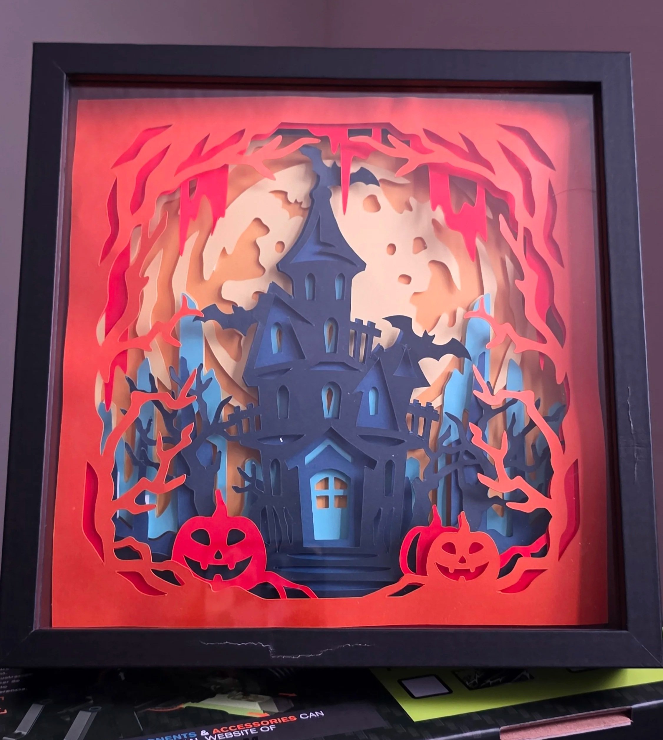 Framed Halloween-themed paper art featuring a haunted house silhouette surrounded by orange and red paper flames and pumpkins, with a purple and black frame.