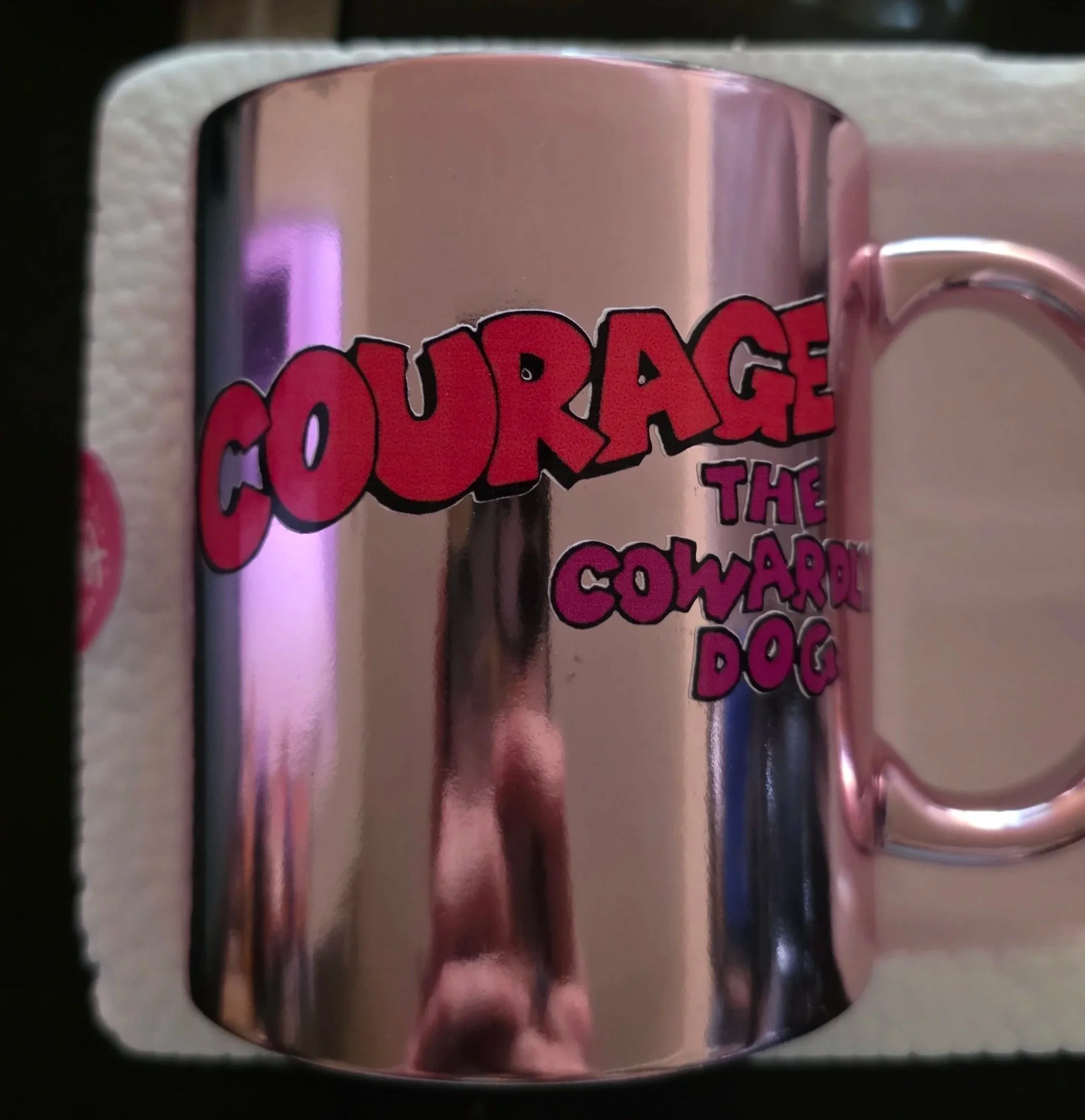 A pink mug with the words "Courage The Cowardly Dog" in red and purple letters. The mug is on a white foam tray with a bottle of water and a ruler nearby.