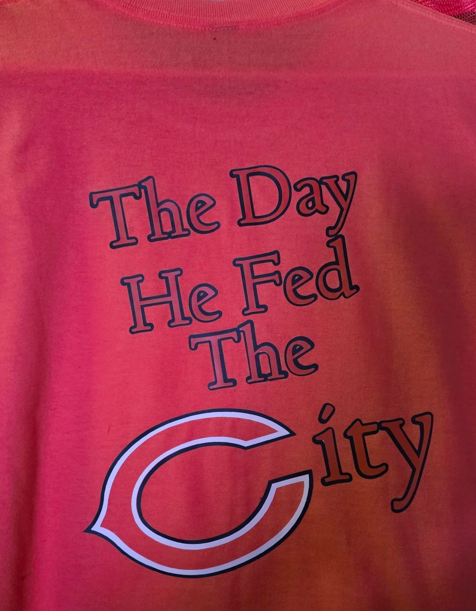 A red T-shirt hanging on a hanger with text that reads, 'The Day He Fed The City,' and a large letter 'C' logo.