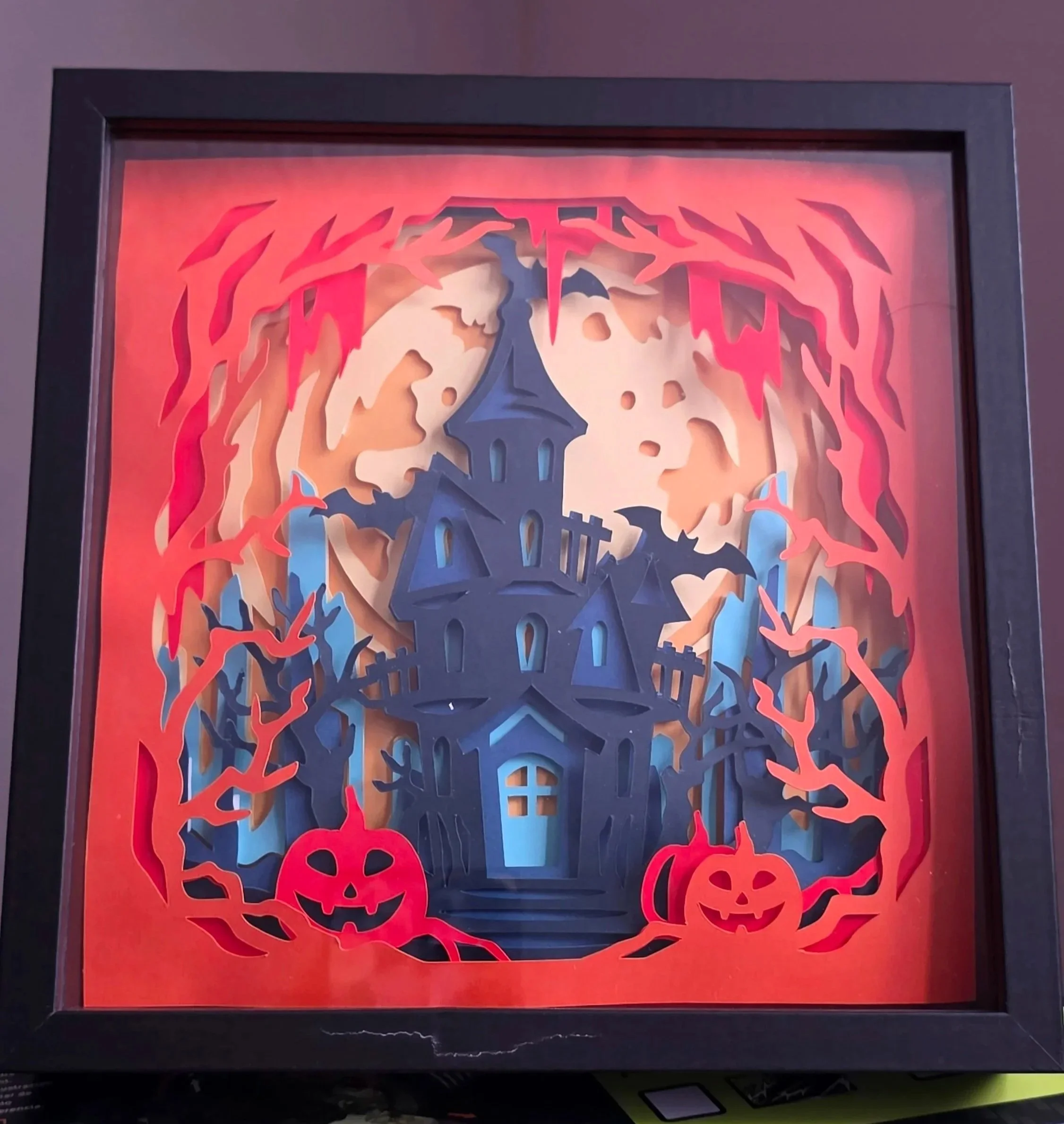 Framed Halloween-themed paper art featuring a haunted house silhouette surrounded by orange and red paper flames and pumpkins, with a purple and black frame.