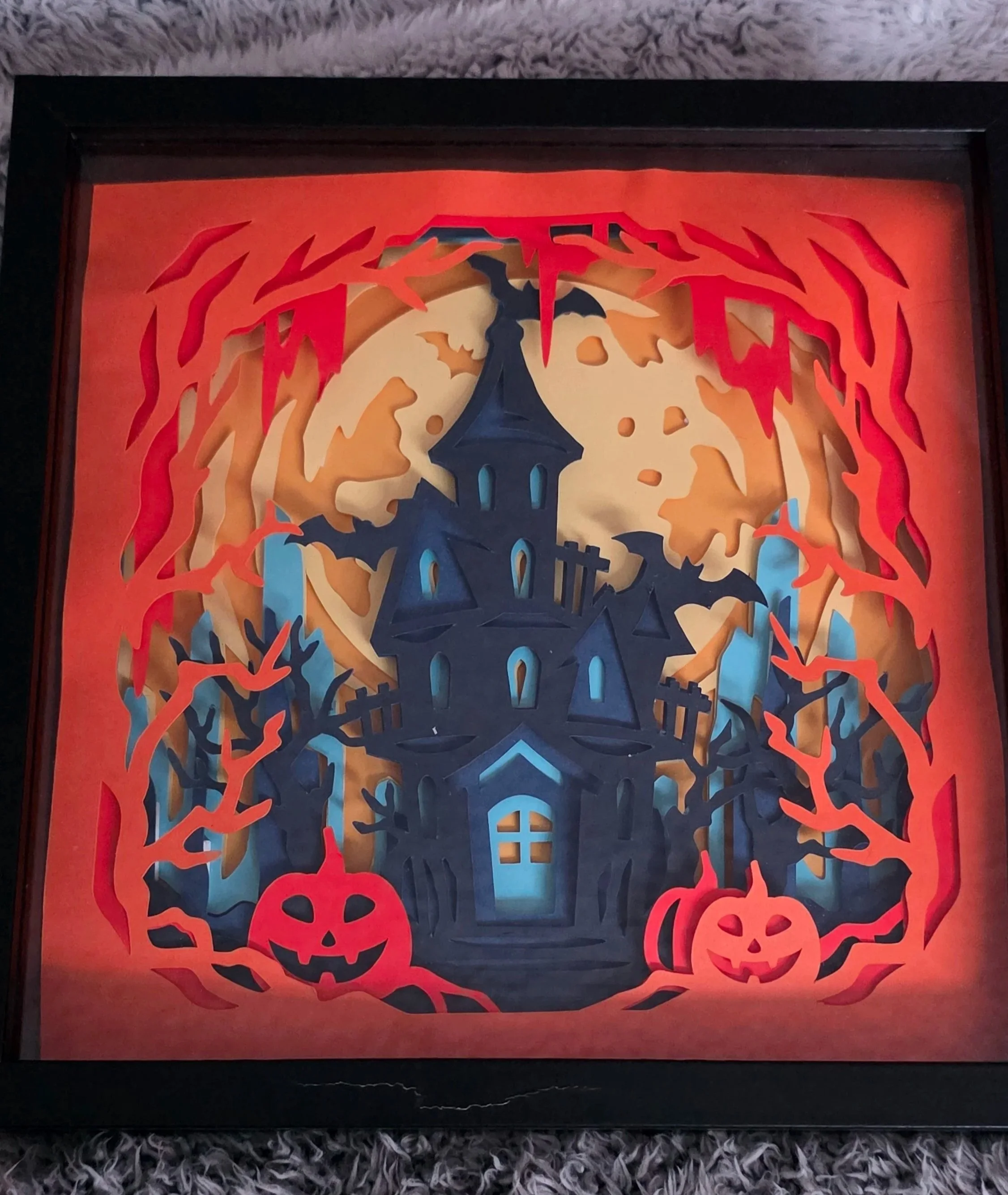 A Halloween-themed paper cutout artwork of a haunted house with a full moon, surrounded by red flames, purple trees, and two smiling pumpkin jack-o'-lanterns at the bottom.