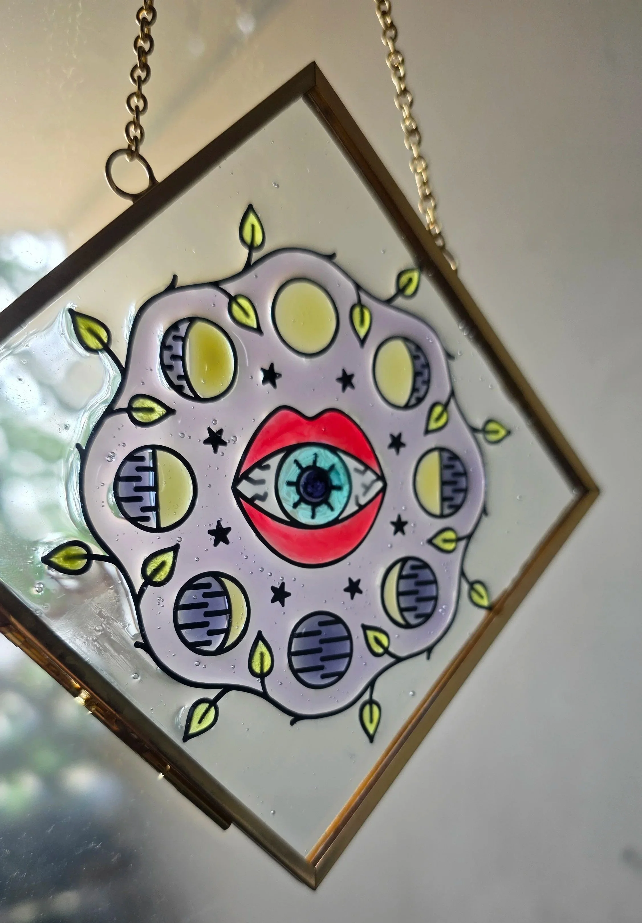 Decorative stained glass piece featuring an eye in the center with red lips, surrounded by moon phases and plant-like details, hanging from a gold chain.