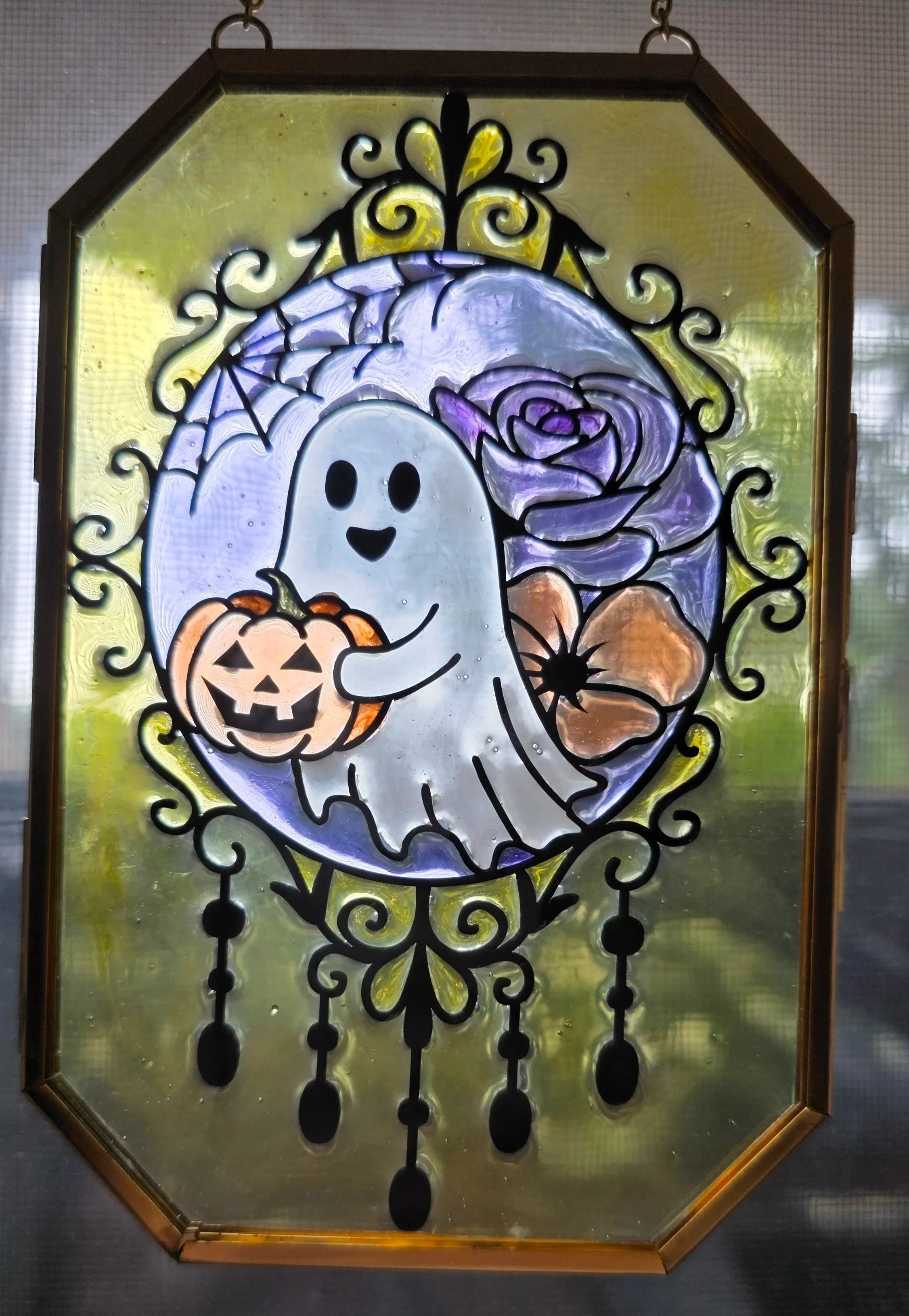 A decorative stained glass window featuring a cartoon ghost holding a carved pumpkin, with purple roses, a spider web, and hanging black chains with spider accents.