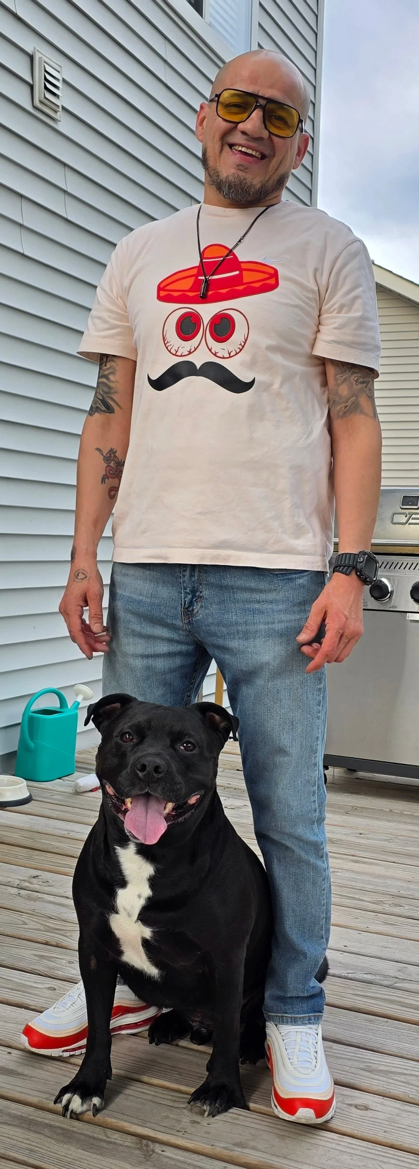 A man wearing sunglasses, a graphic t-shirt with cartoon eyes, a hat, and a mustache design, standing outside with a black dog sitting in front of him on a wooden deck.